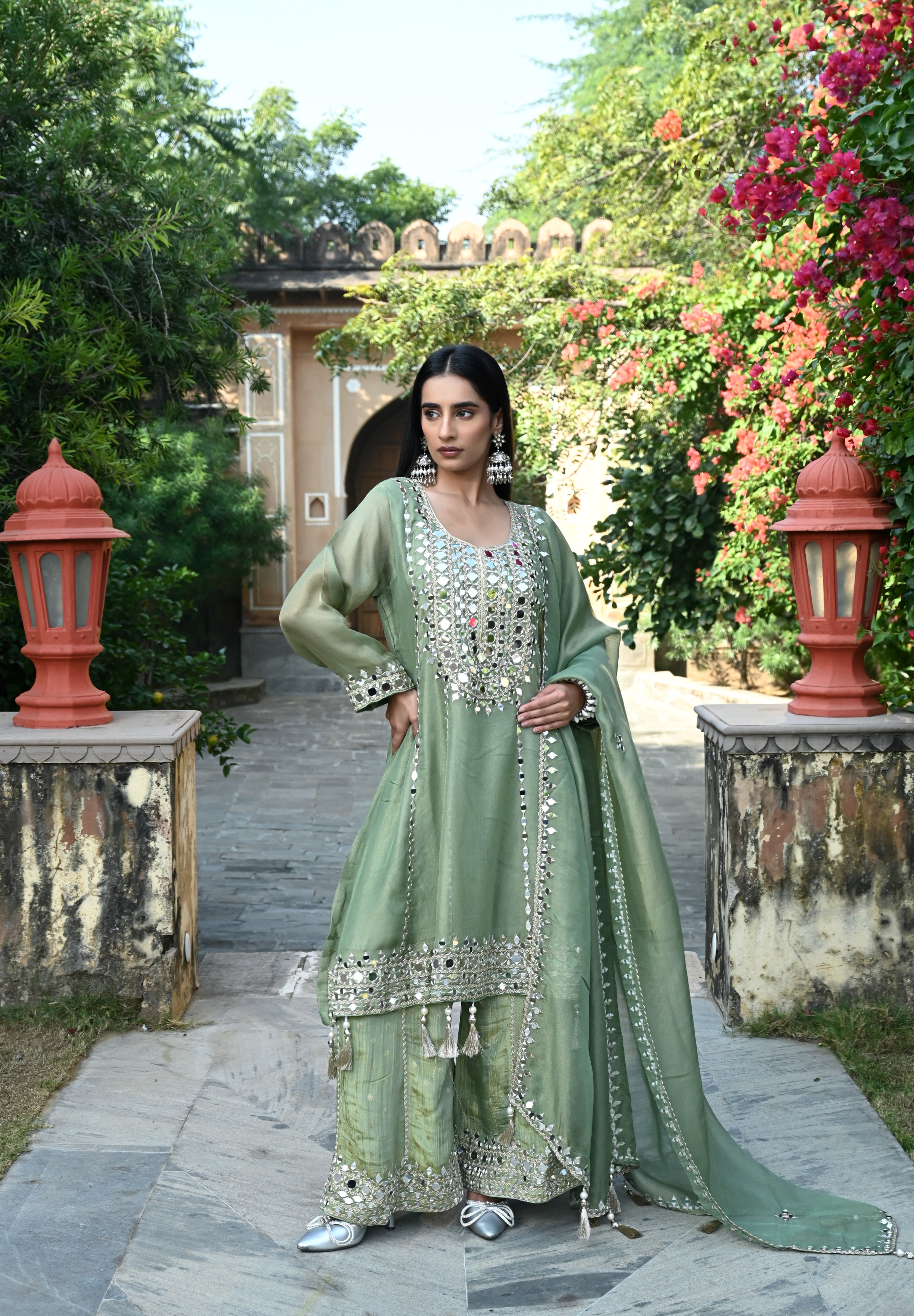 Sage-green traditional outfit featuring a mirror-embroidered kurta with wide sleeves, matching wide-legged trousers with embellished hems, and a coordinating dupatta finished with reflective detailing and tassels.