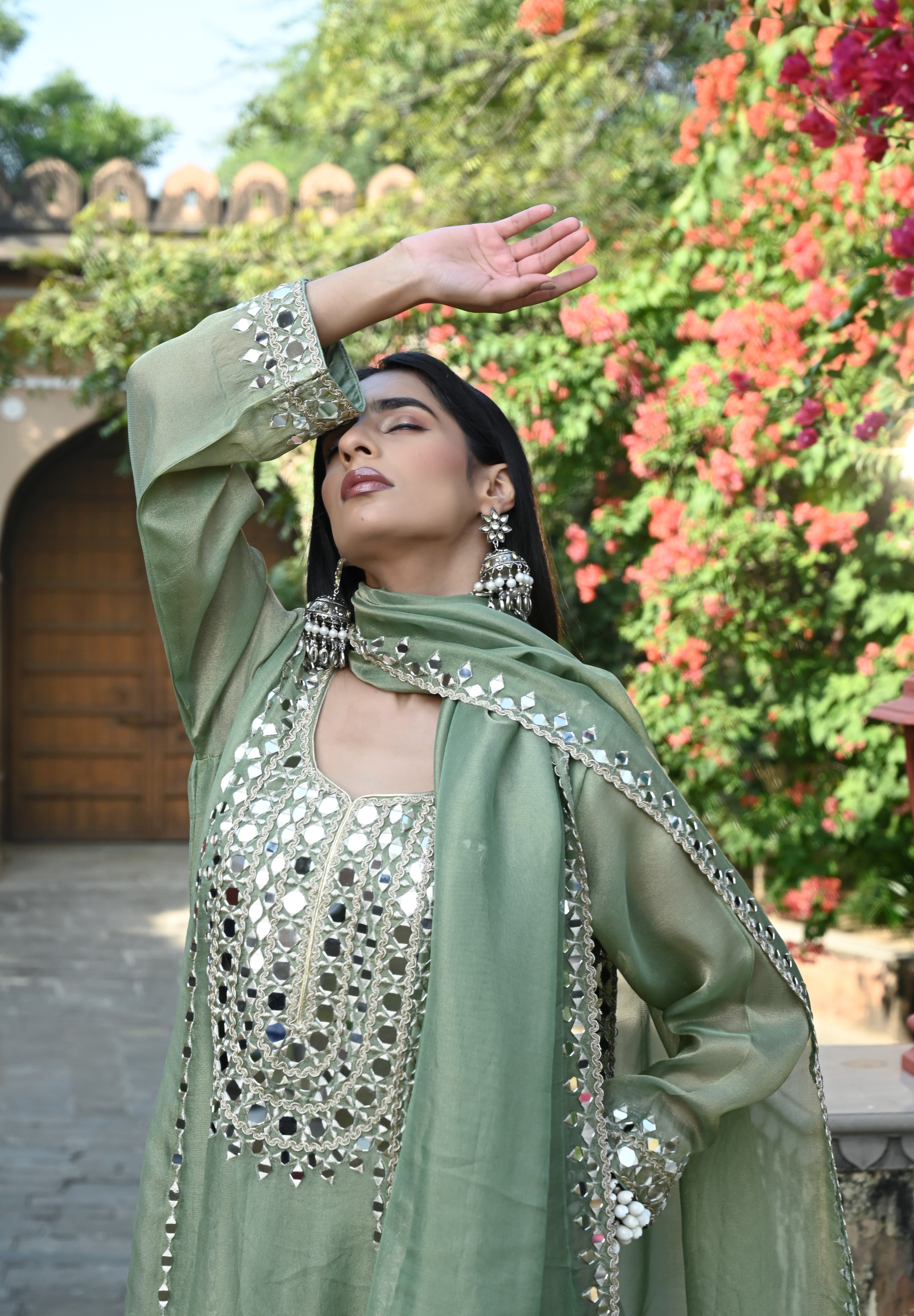 Sage-green traditional outfit featuring a mirror-embroidered kurta with wide sleeves, matching wide-legged trousers with embellished hems, and a coordinating dupatta finished with reflective detailing and tassels.