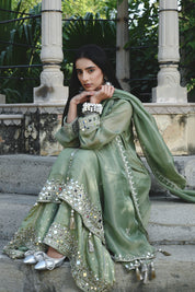 Sage-green traditional outfit featuring a mirror-embroidered kurta with wide sleeves, matching wide-legged trousers with embellished hems, and a coordinating dupatta finished with reflective detailing and tassels.