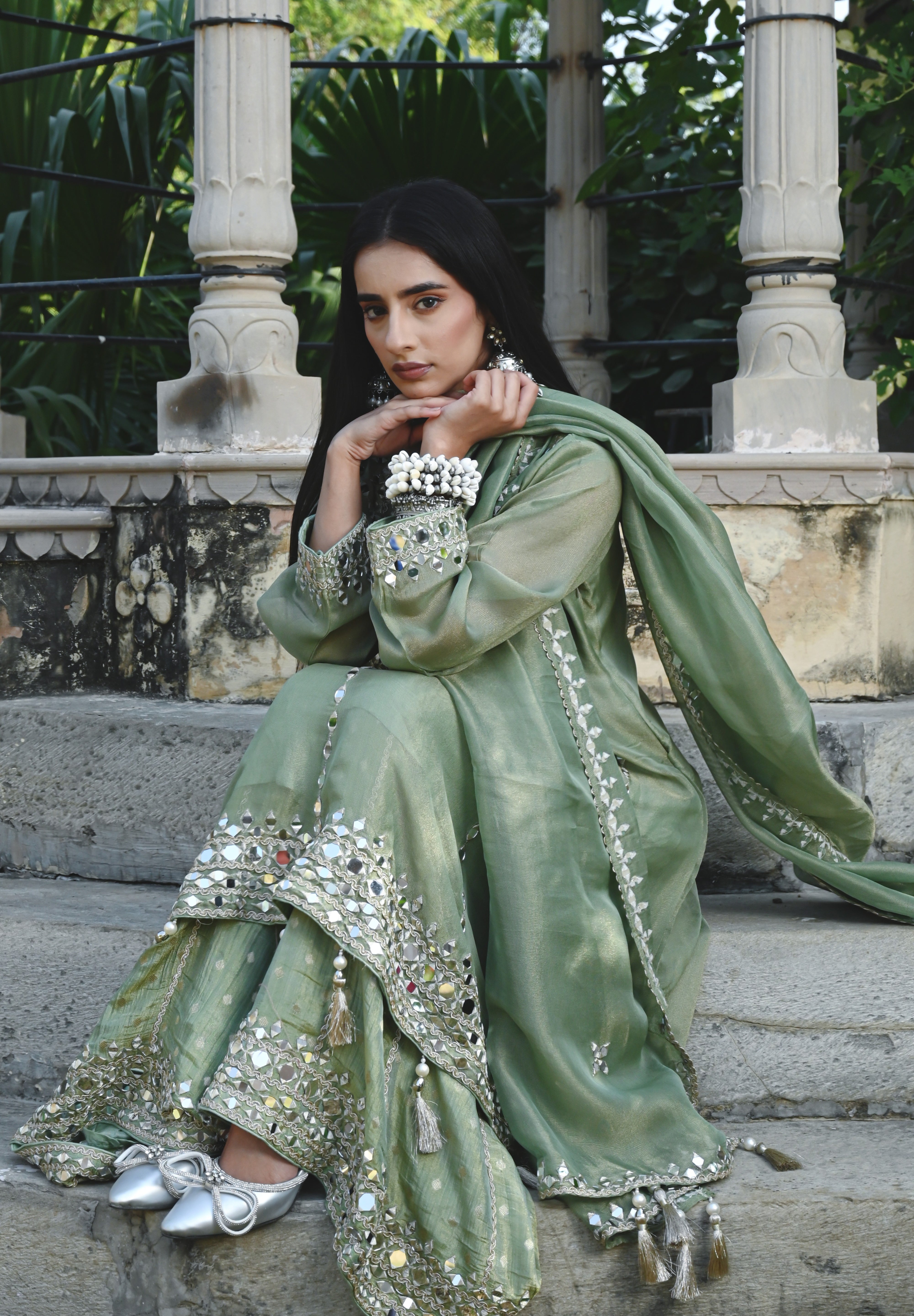 Sage-green traditional outfit featuring a mirror-embroidered kurta with wide sleeves, matching wide-legged trousers with embellished hems, and a coordinating dupatta finished with reflective detailing and tassels.