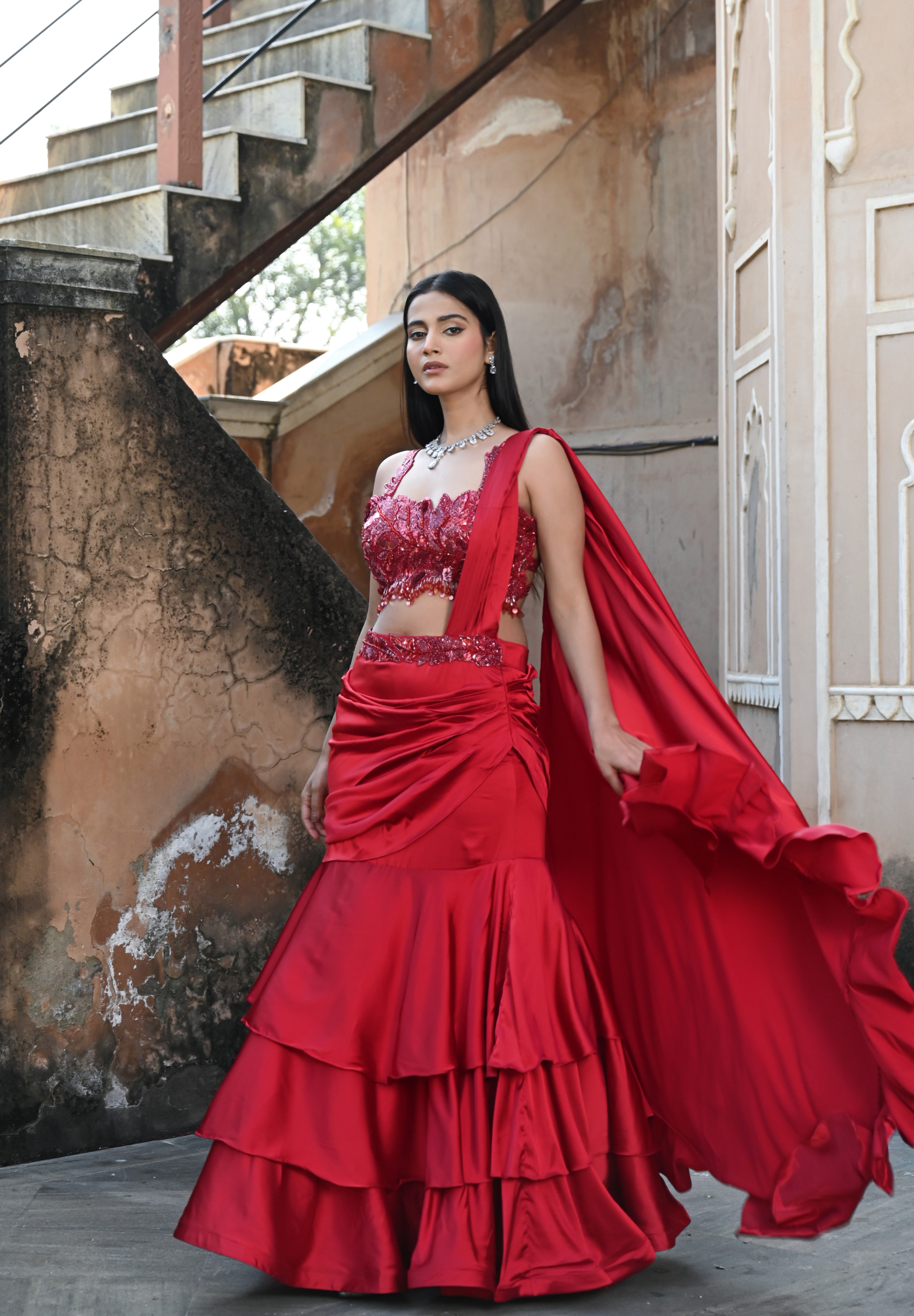 Model wearing a deep red embellished lehenga set with a sequined blouse, layered flared skirt, and flowing cape-style dupatta, styled with statement jewellery, posed in a heritage courtyard for Tulsi Studio Lifestyle wedding collection.