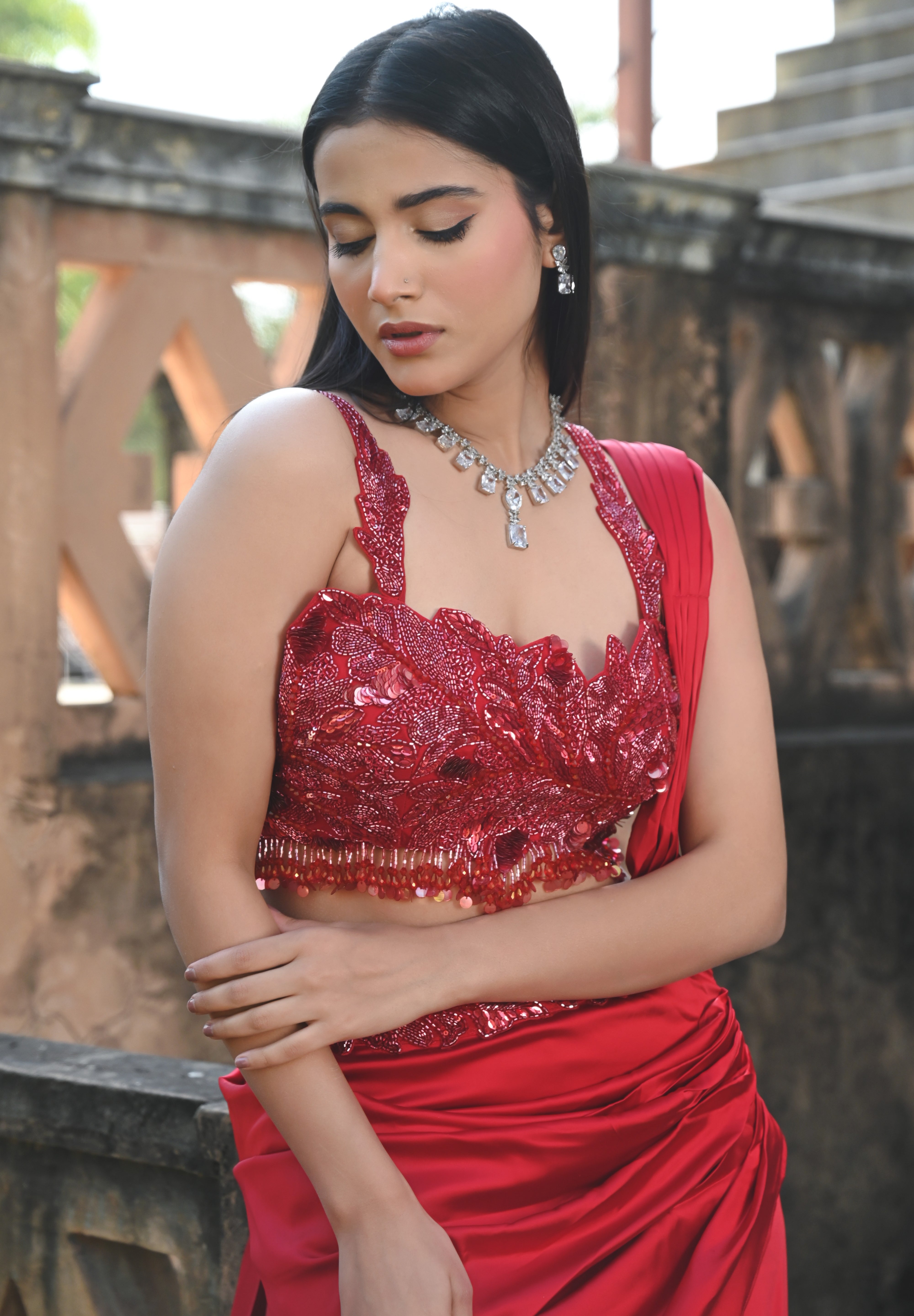 Model wearing a deep red embellished lehenga set with a sequined blouse, layered flared skirt, and flowing cape-style dupatta, styled with statement jewellery, posed in a heritage courtyard for Tulsi Studio Lifestyle wedding collection.