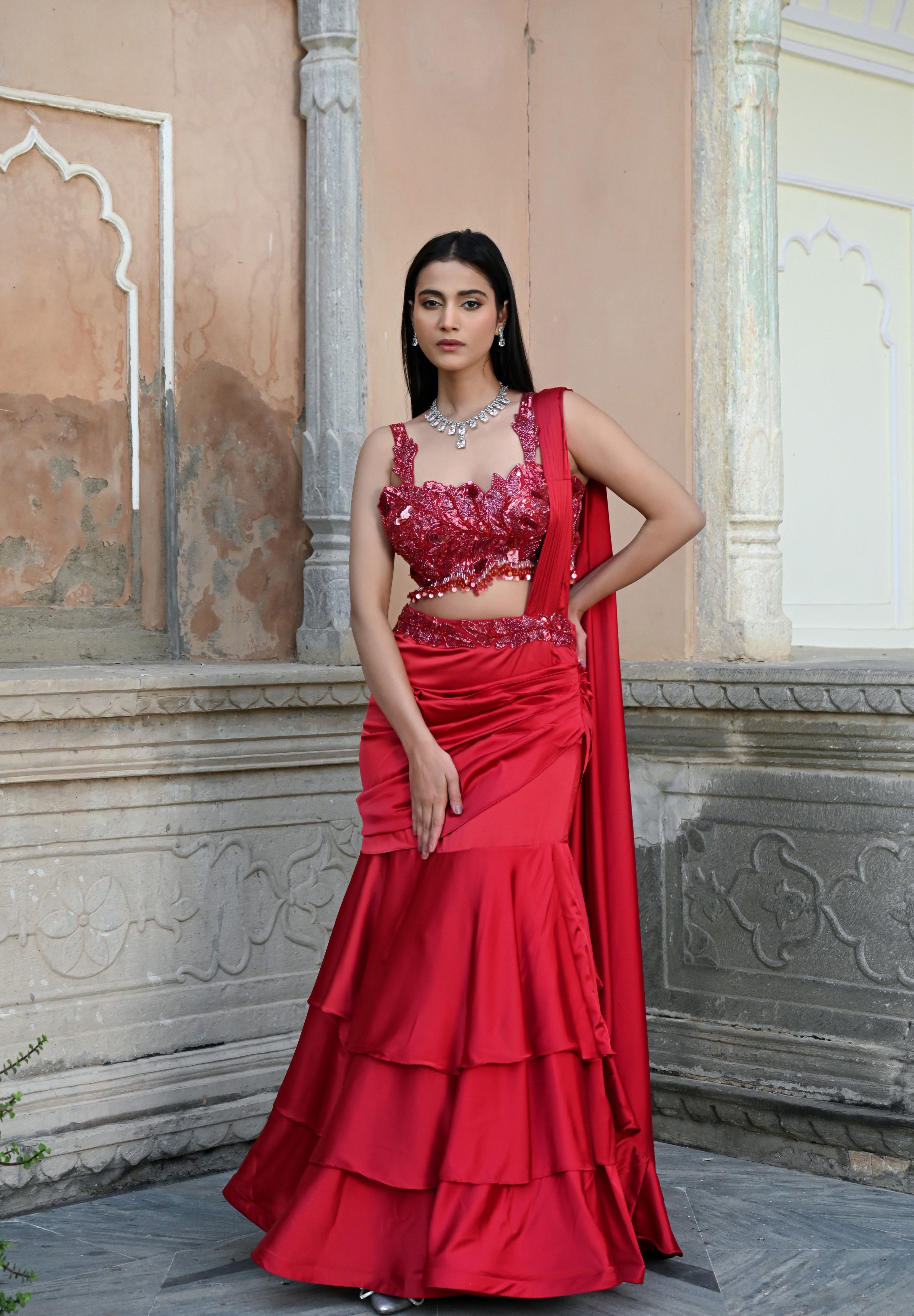 Model wearing a deep red embellished lehenga set with a sequined blouse, layered flared skirt, and flowing cape-style dupatta, styled with statement jewellery, posed in a heritage courtyard for Tulsi Studio Lifestyle wedding collection.