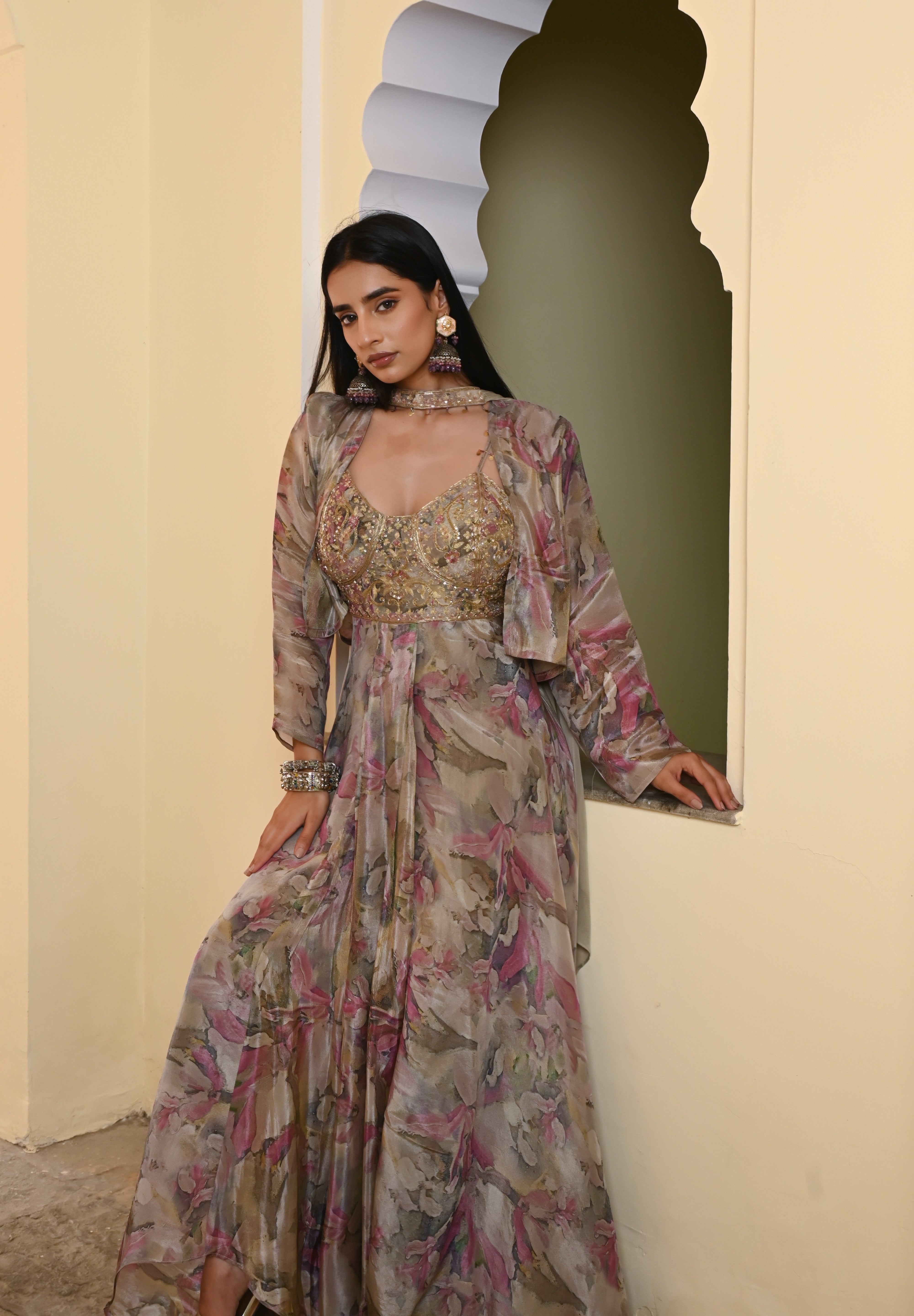 Model wearing a pastel floral printed Anarkali gown with an embroidered bustier and sheer jacket, styled with statement earrings and bangles, posing against a heritage archway for Tulsi Studio festive collection.