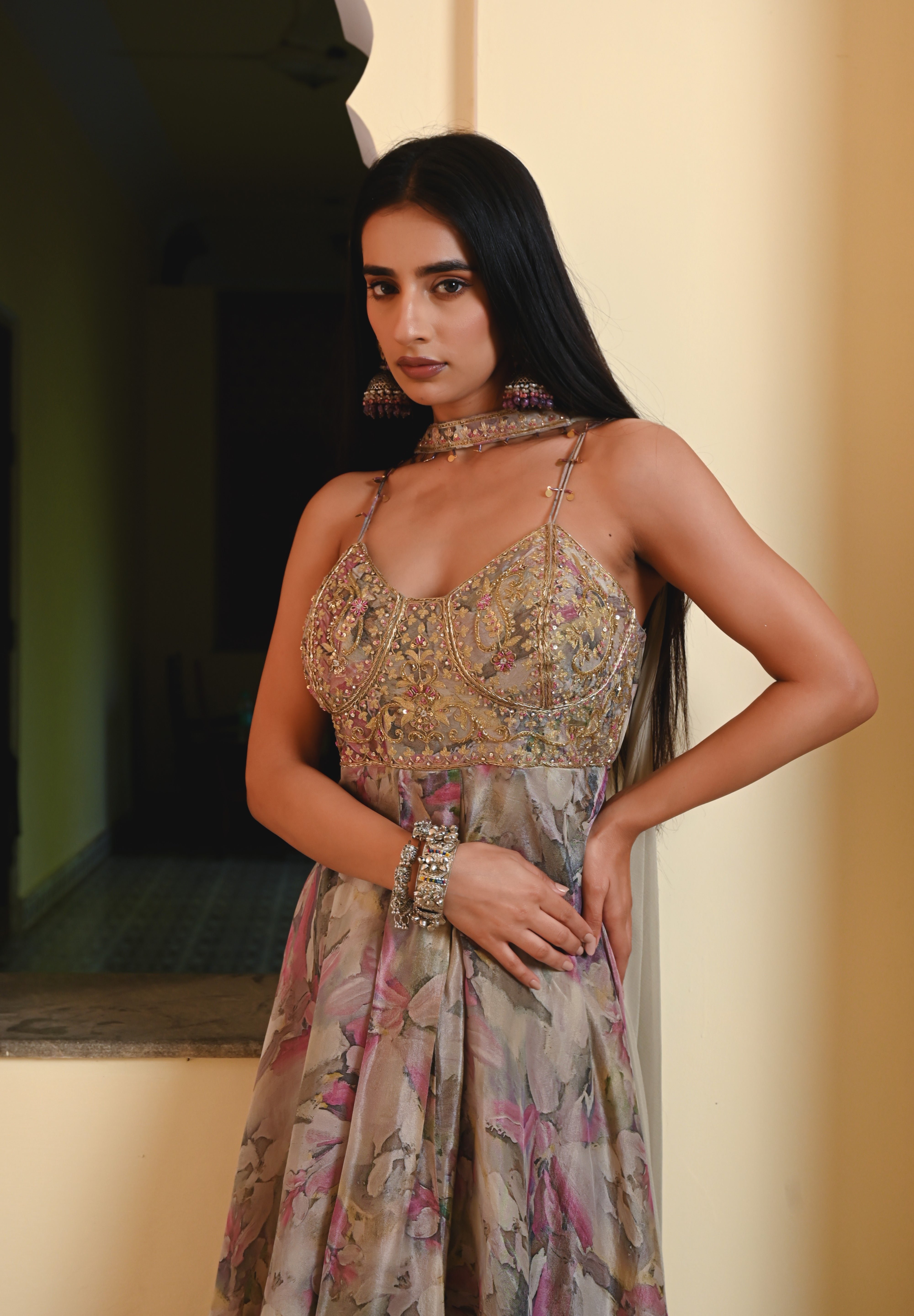 Model wearing a pastel floral printed Anarkali gown with an embroidered bustier and sheer jacket, styled with statement earrings and bangles, posing against a heritage archway for Tulsi Studio festive collection.