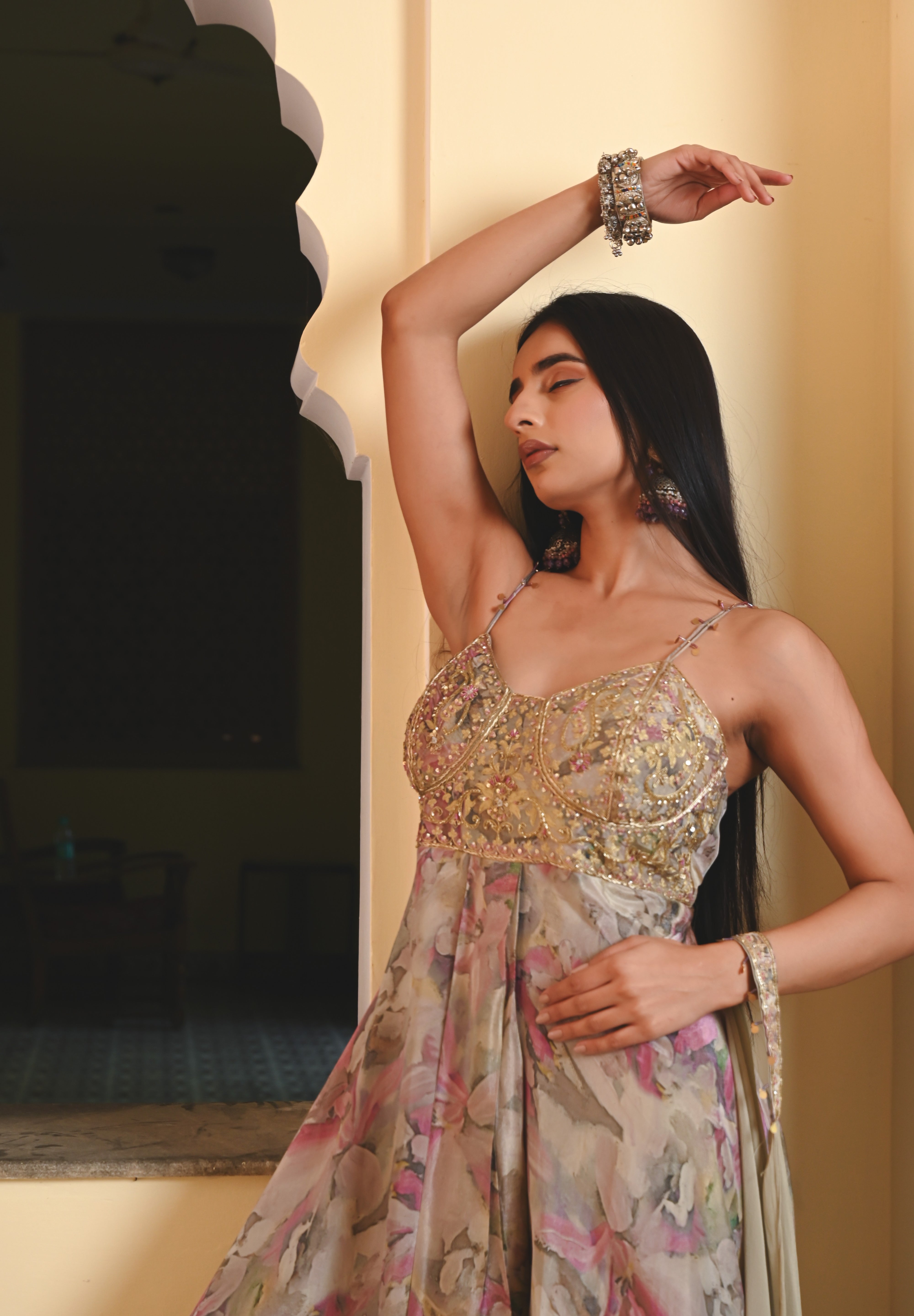 Model wearing a pastel floral printed Anarkali gown with an embroidered bustier and sheer jacket, styled with statement earrings and bangles, posing against a heritage archway for Tulsi Studio festive collection.