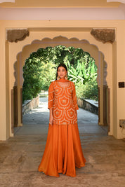 Model wearing an orange embroidered kurta with intricate threadwork paired with flared sharara pants, styled with statement earrings, standing under a heritage archway surrounded by lush greenery for Tulsi Studio festive collection.