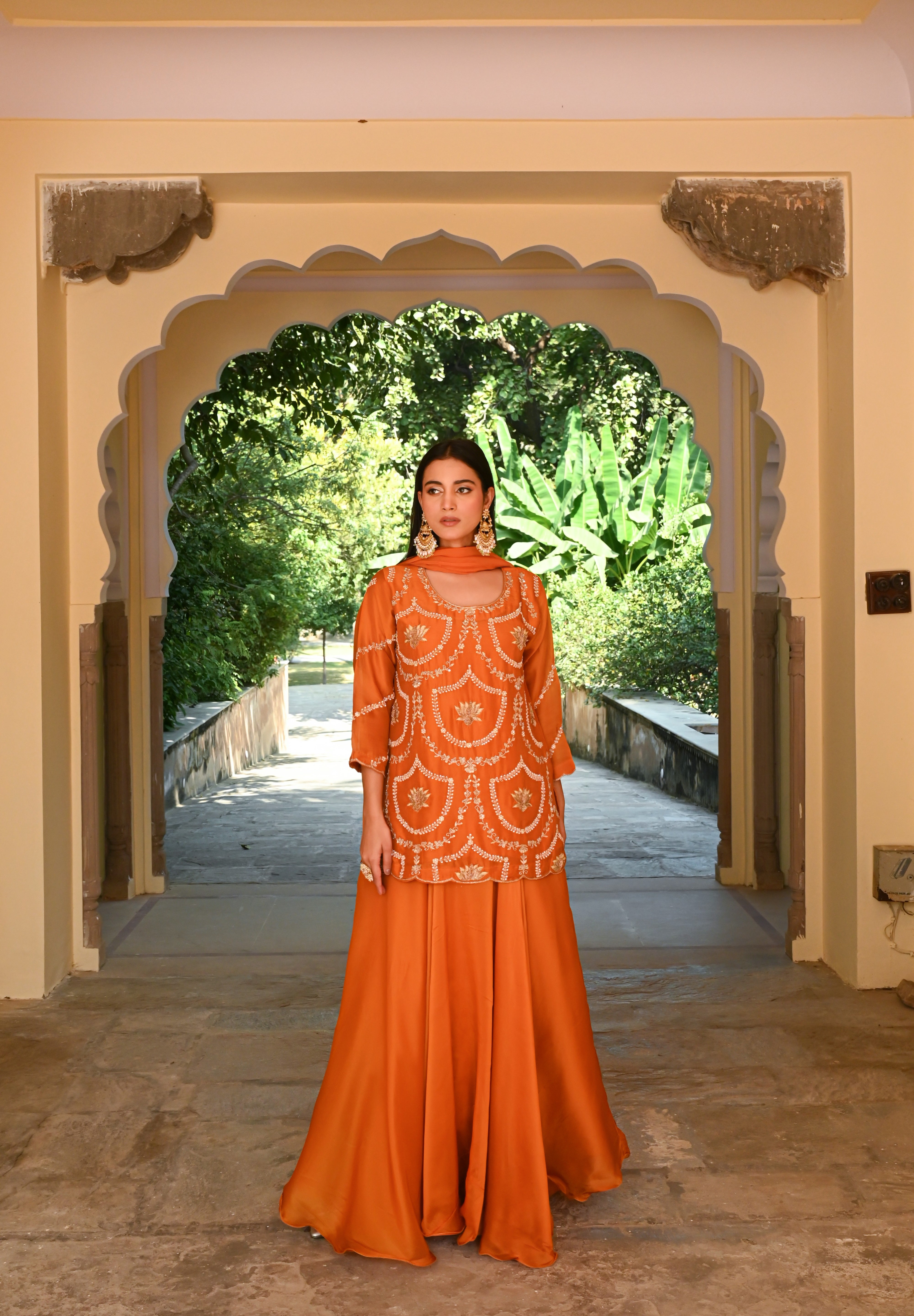 Model wearing an orange embroidered kurta with intricate threadwork paired with flared sharara pants, styled with statement earrings, standing under a heritage archway surrounded by lush greenery for Tulsi Studio festive collection.