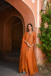 Model wearing an orange embroidered kurta with intricate threadwork paired with flared sharara pants, styled with statement earrings, standing under a heritage archway surrounded by lush greenery for Tulsi Studio festive collection.