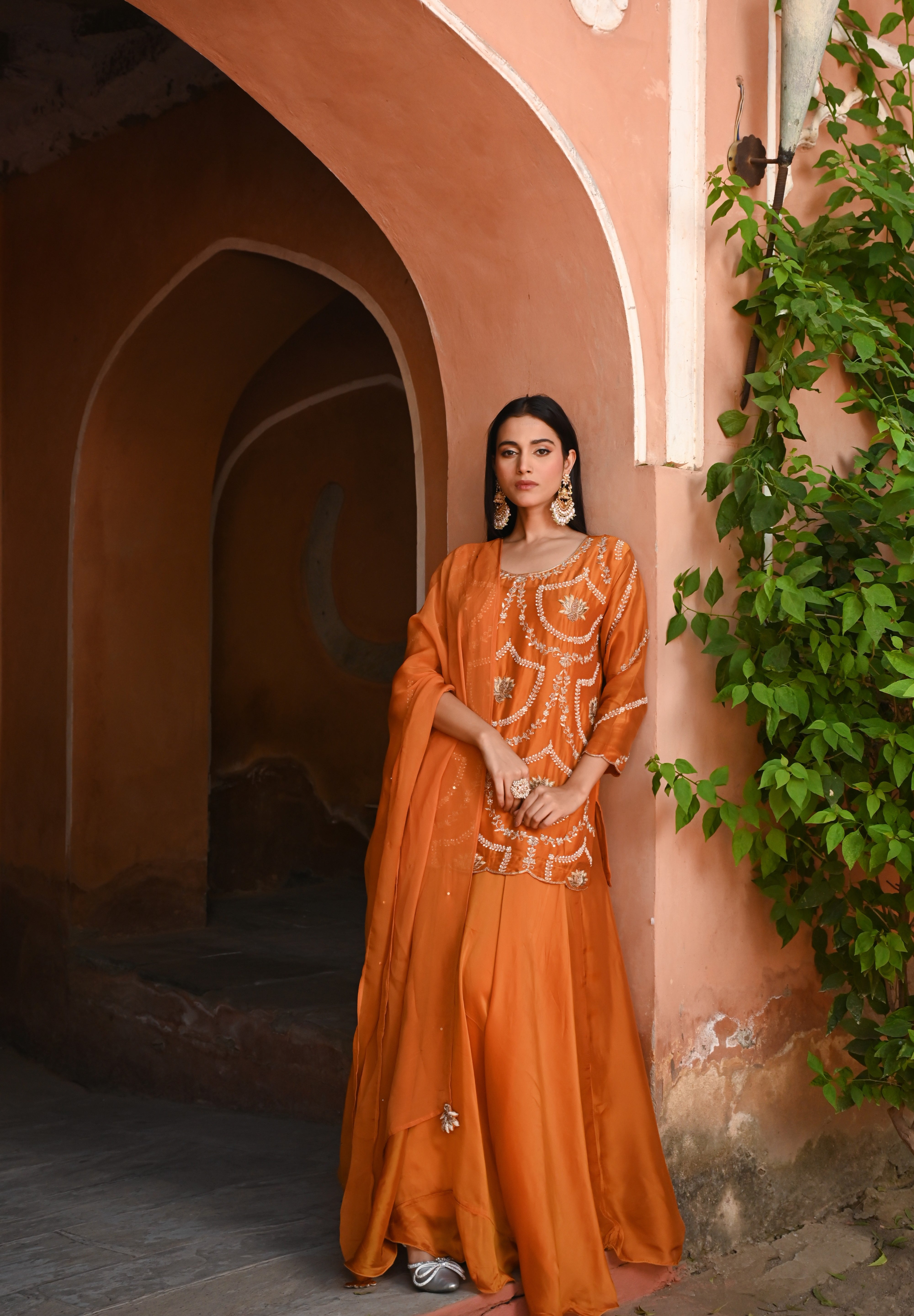 Model wearing an orange embroidered kurta with intricate threadwork paired with flared sharara pants, styled with statement earrings, standing under a heritage archway surrounded by lush greenery for Tulsi Studio festive collection.