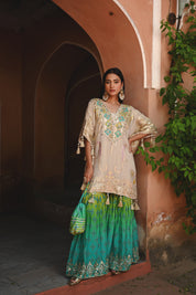 Model wearing a beige embroidered kurta with gold and turquoise detailing paired with a teal green sharara, styled with statement earrings and potli bag, posing in a heritage archway for Tulsi Studio festive wedding collection.