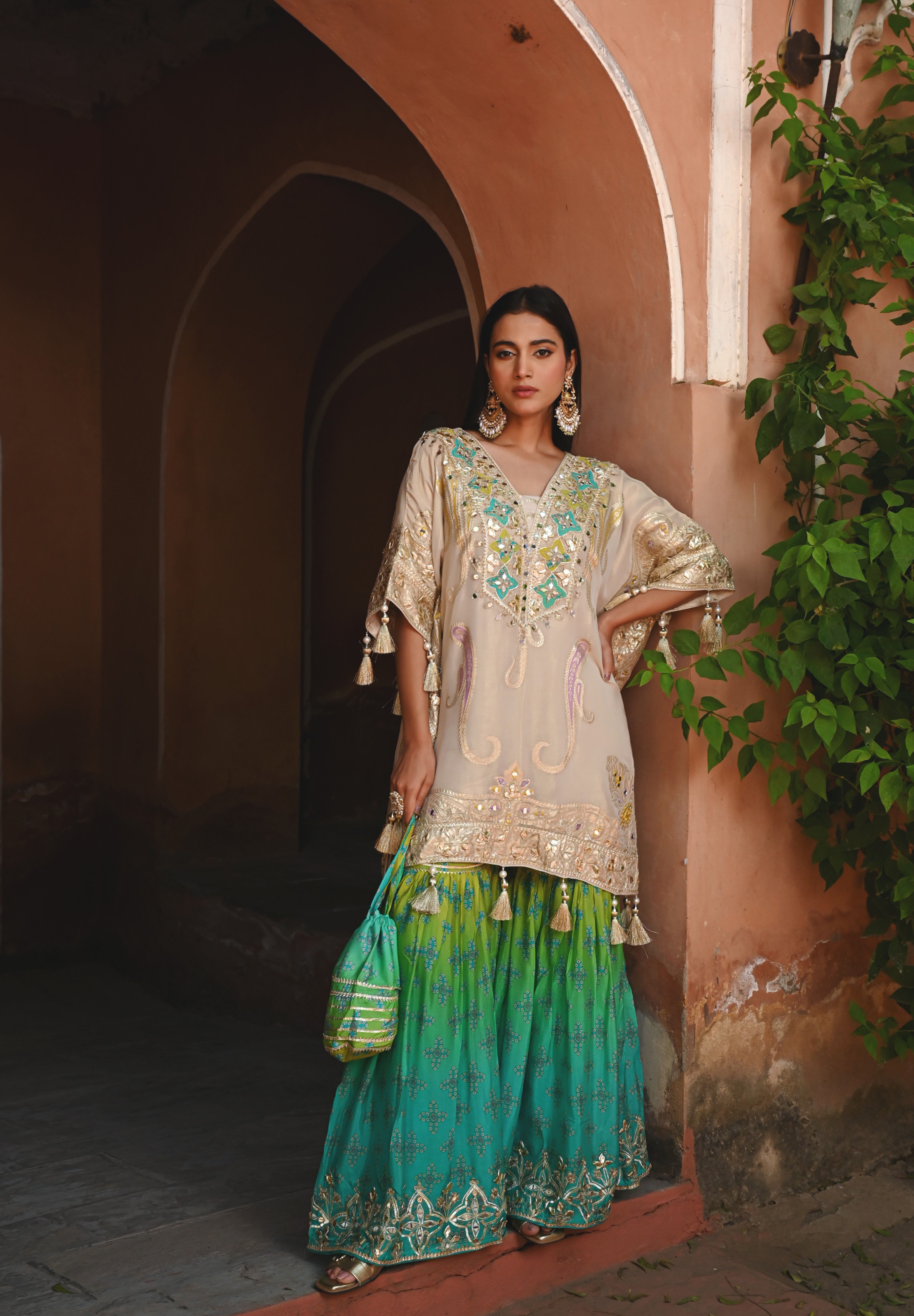 Model wearing a beige embroidered kurta with gold and turquoise detailing paired with a teal green sharara, styled with statement earrings and potli bag, posing in a heritage archway for Tulsi Studio festive wedding collection.