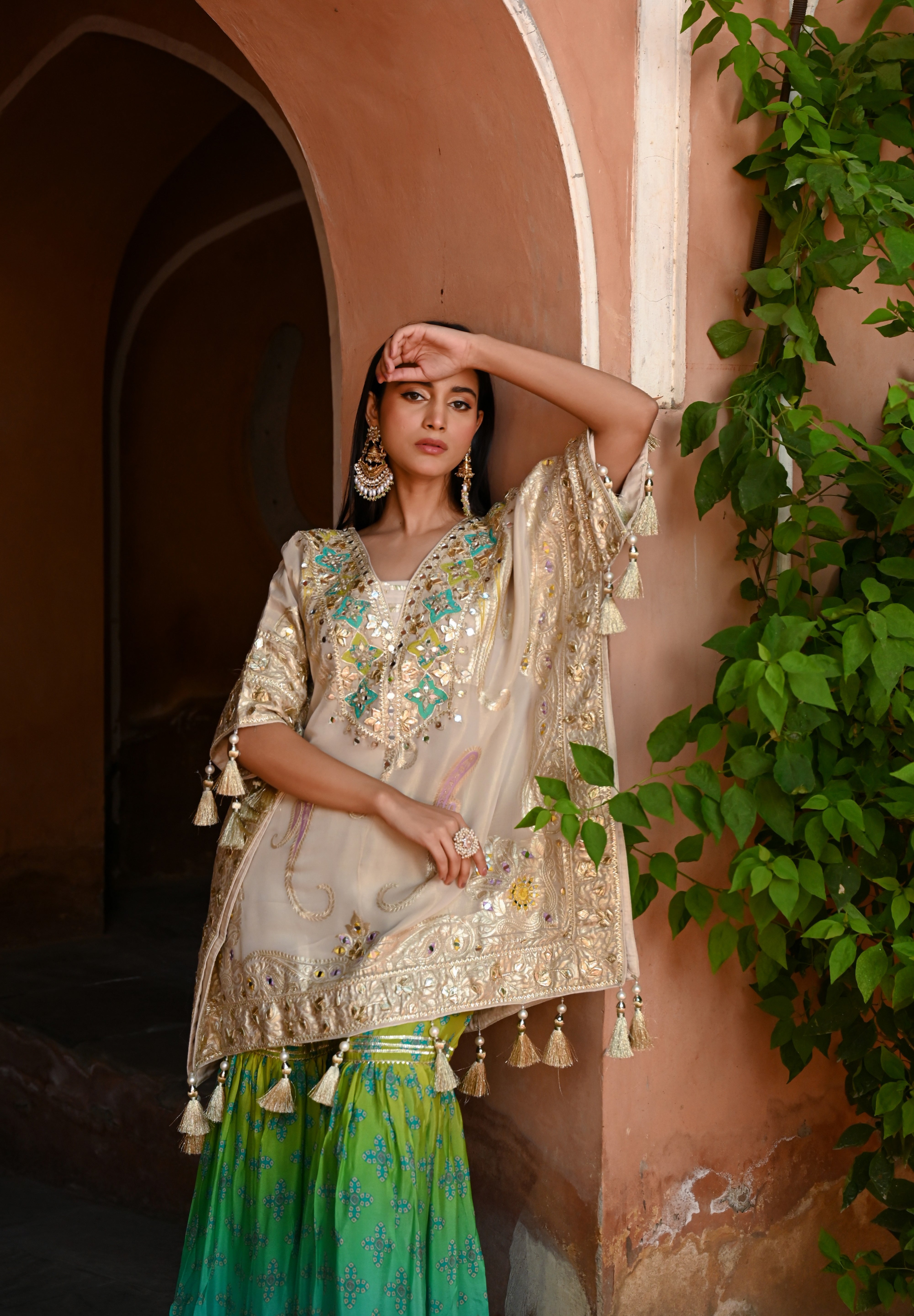 Model wearing a beige embroidered kurta with gold and turquoise detailing paired with a teal green sharara, styled with statement earrings and potli bag, posing in a heritage archway for Tulsi Studio festive wedding collection.