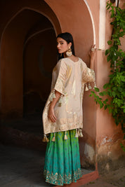 Model wearing a beige embroidered kurta with gold and turquoise detailing paired with a teal green sharara, styled with statement earrings and potli bag, posing in a heritage archway for Tulsi Studio festive wedding collection.
