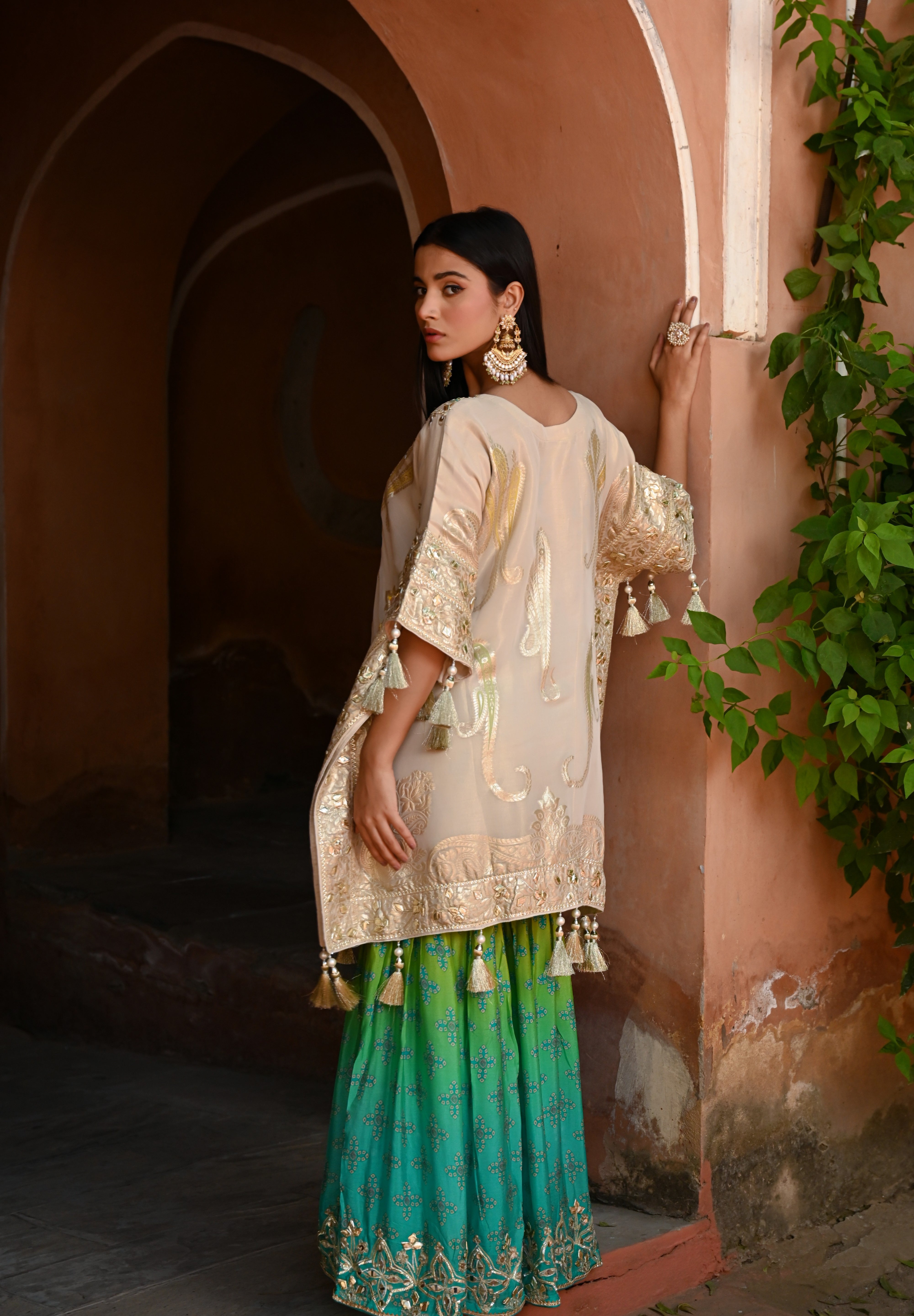 Model wearing a beige embroidered kurta with gold and turquoise detailing paired with a teal green sharara, styled with statement earrings and potli bag, posing in a heritage archway for Tulsi Studio festive wedding collection.