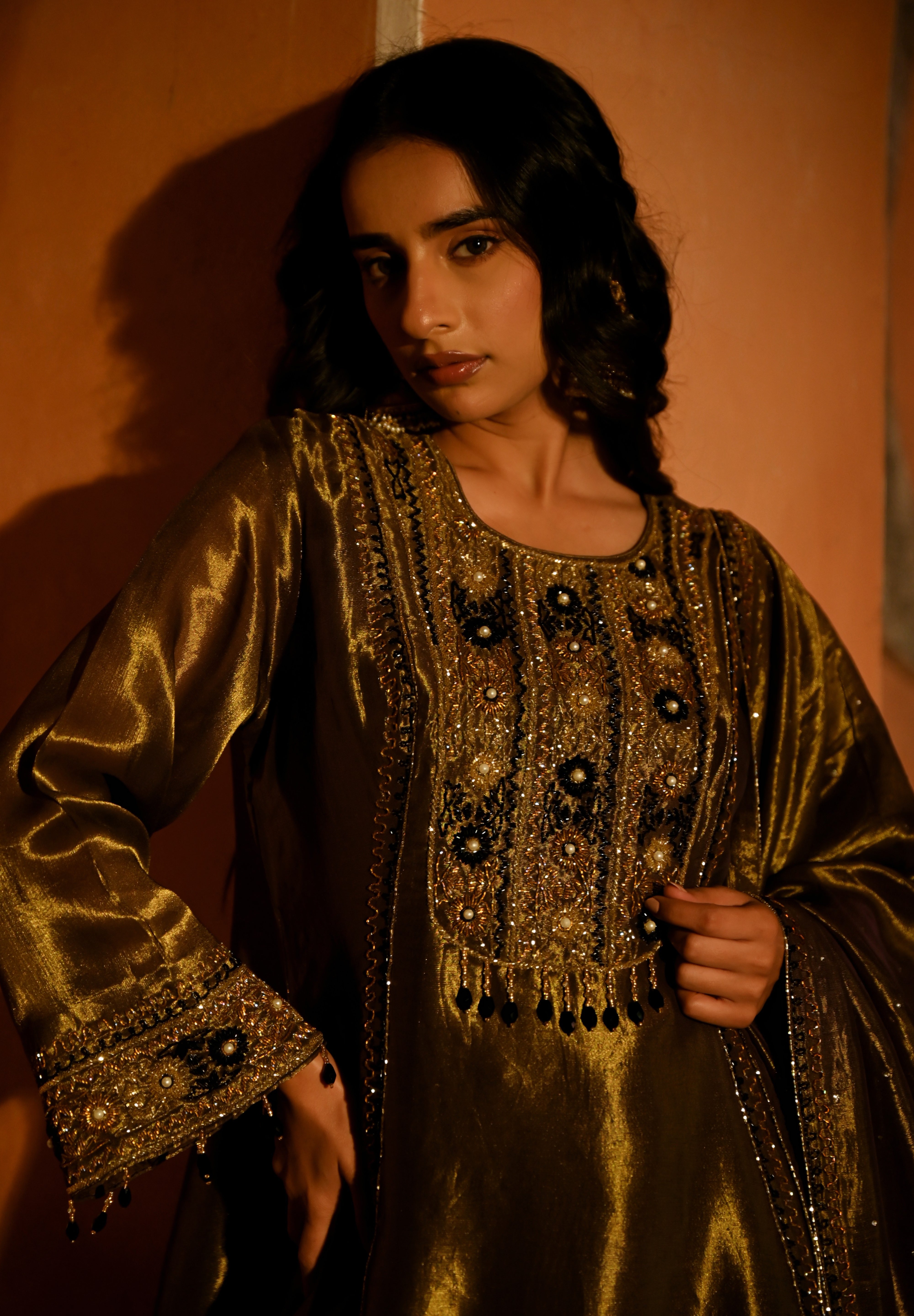 Woman in a gold embroidered traditional outfit against an orange wall.