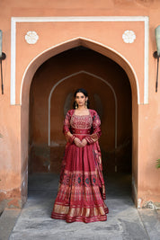 Model wearing a deep maroon embroidered Anarkali suit with gold detailing and printed dupatta, styled with traditional jhumka earrings, posing against an earthy arched heritage backdrop for Tulsi Studio wedding collection.
