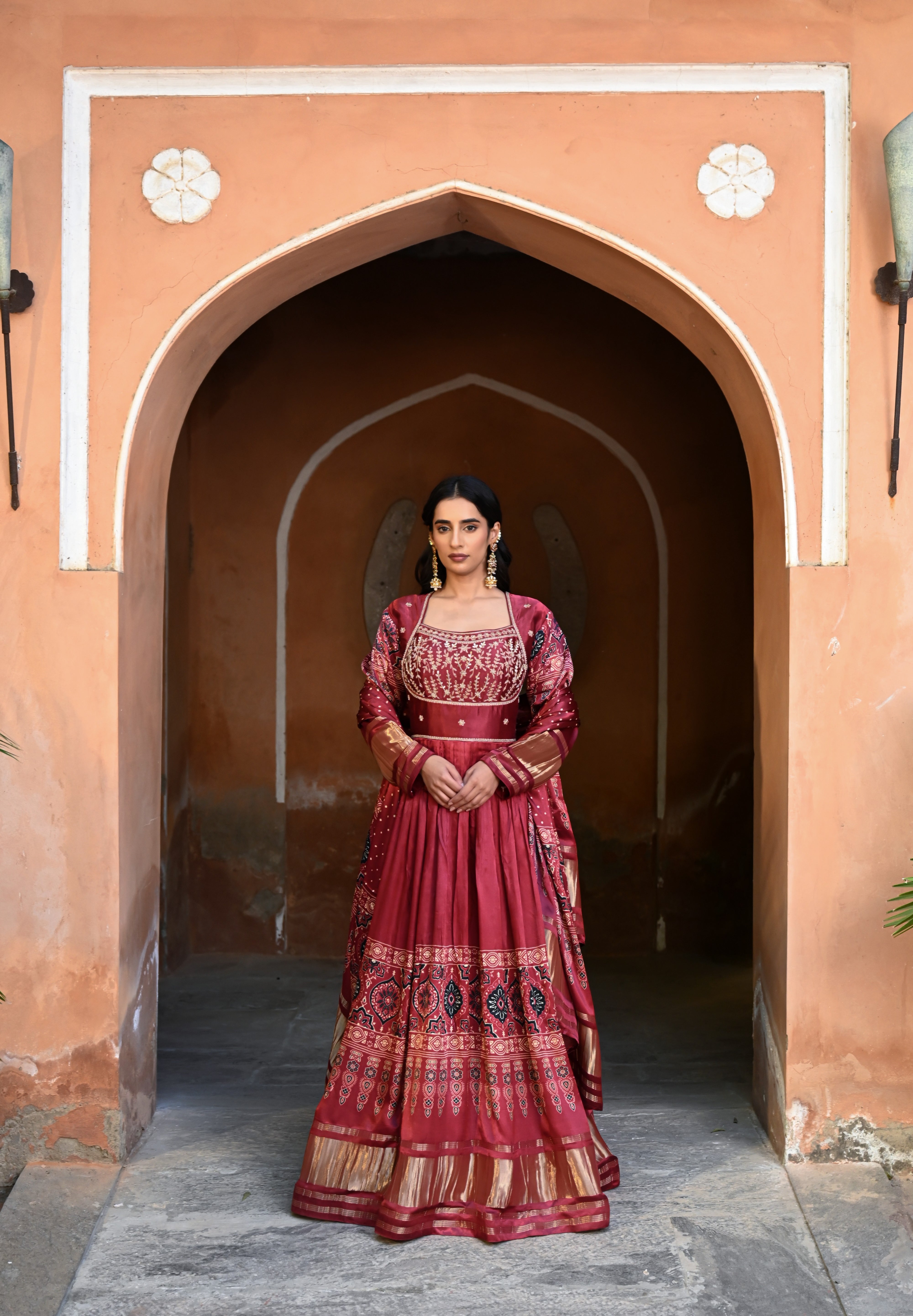 Model wearing a deep maroon embroidered Anarkali suit with gold detailing and printed dupatta, styled with traditional jhumka earrings, posing against an earthy arched heritage backdrop for Tulsi Studio wedding collection.
