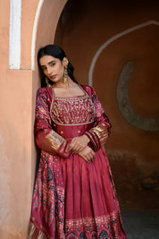 Model wearing a deep maroon embroidered Anarkali suit with gold detailing and printed dupatta, styled with traditional jhumka earrings, posing against an earthy arched heritage backdrop for Tulsi Studio wedding collection.

