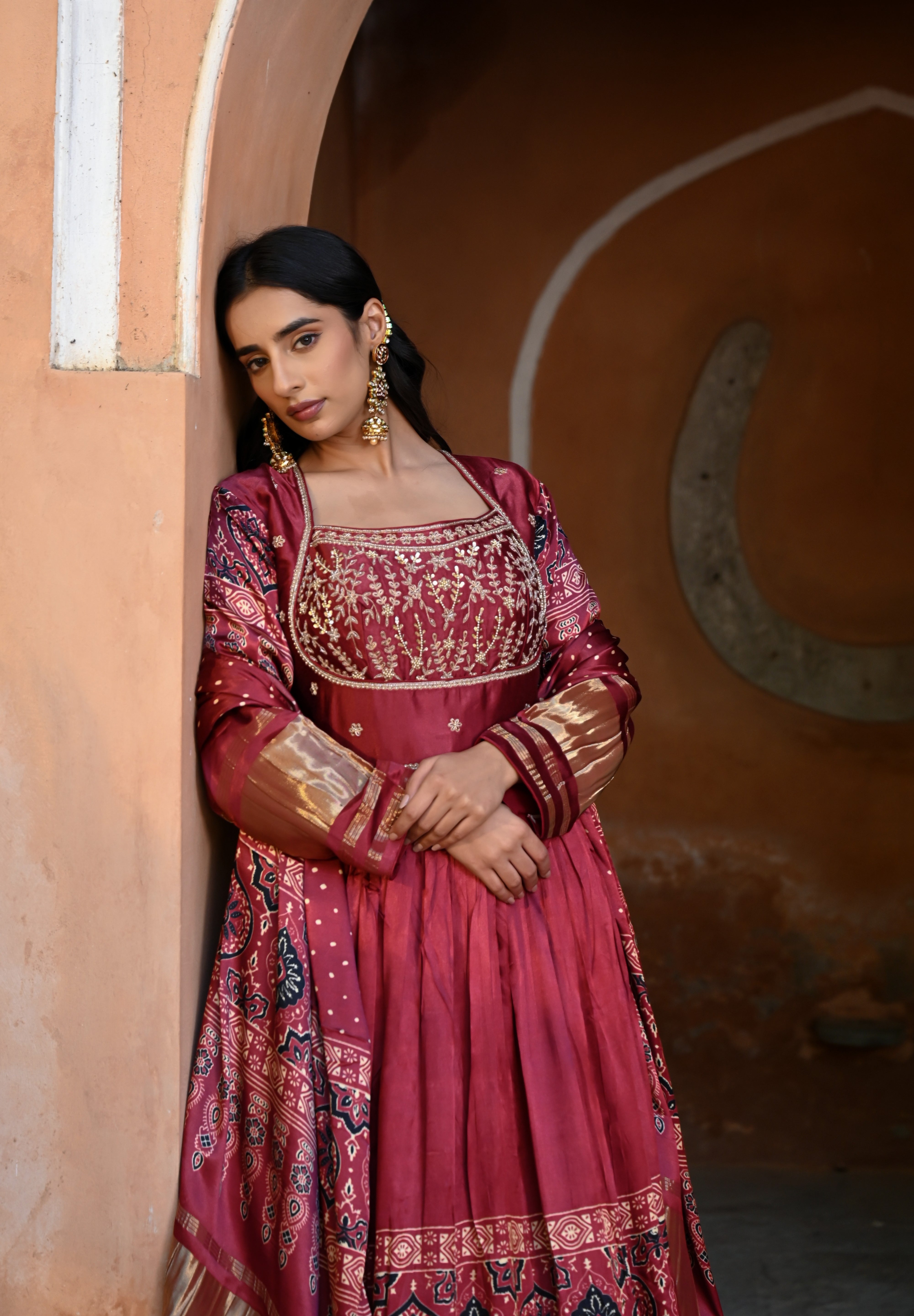 Model wearing a deep maroon embroidered Anarkali suit with gold detailing and printed dupatta, styled with traditional jhumka earrings, posing against an earthy arched heritage backdrop for Tulsi Studio wedding collection.
