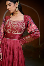 Model wearing a deep maroon embroidered Anarkali suit with gold detailing and printed dupatta, styled with traditional jhumka earrings, posing against an earthy arched heritage backdrop for Tulsi Studio wedding collection.
