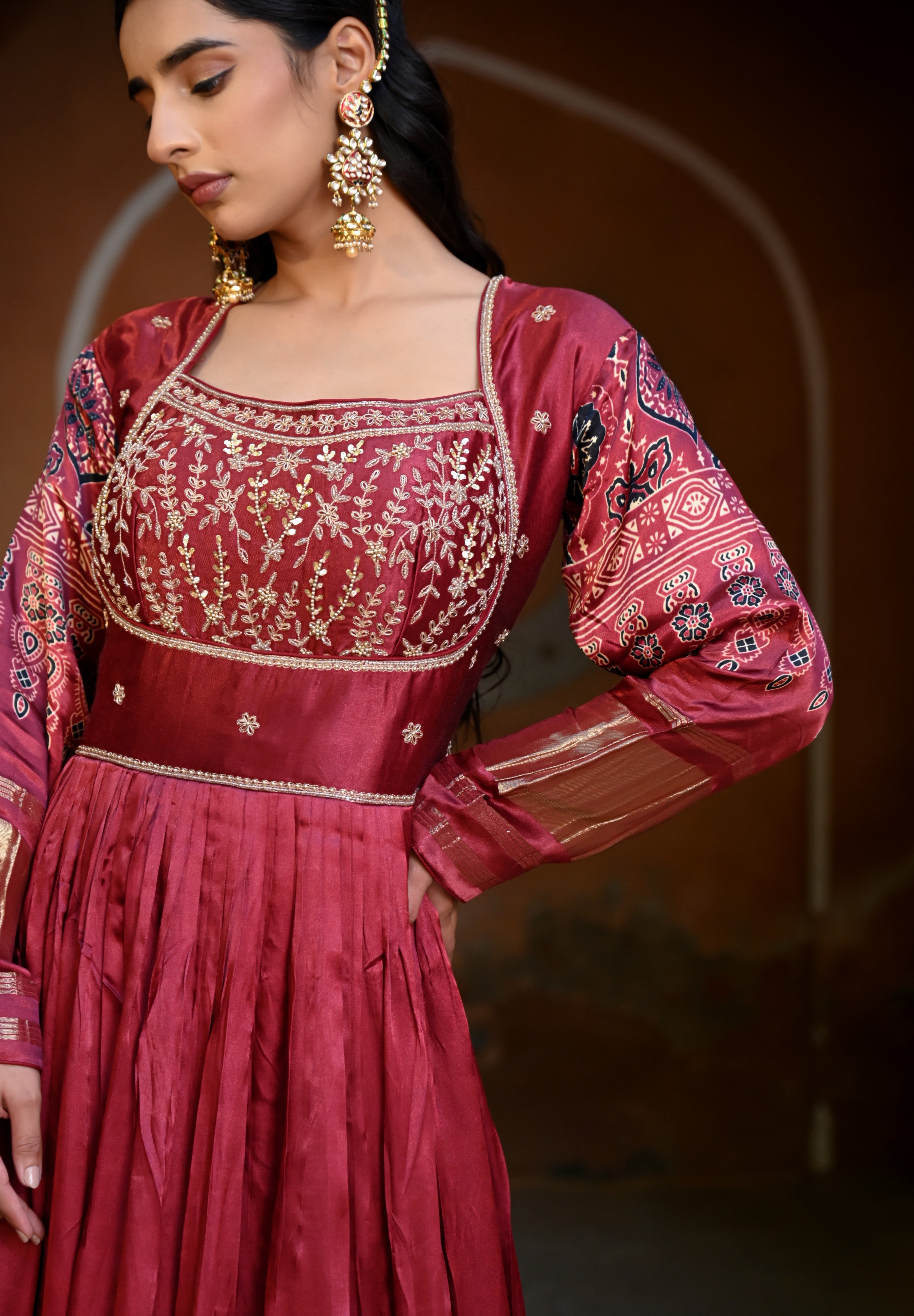 Model wearing a deep maroon embroidered Anarkali suit with gold detailing and printed dupatta, styled with traditional jhumka earrings, posing against an earthy arched heritage backdrop for Tulsi Studio wedding collection.
