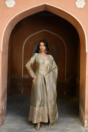 Model wearing an olive-gold embroidered Anarkali dress featuring intricate zari and sequin work, a deep V-neckline, full sleeves, and a rich embellished hem, paired with a matching dupatta and styled under a traditional heritage archway.