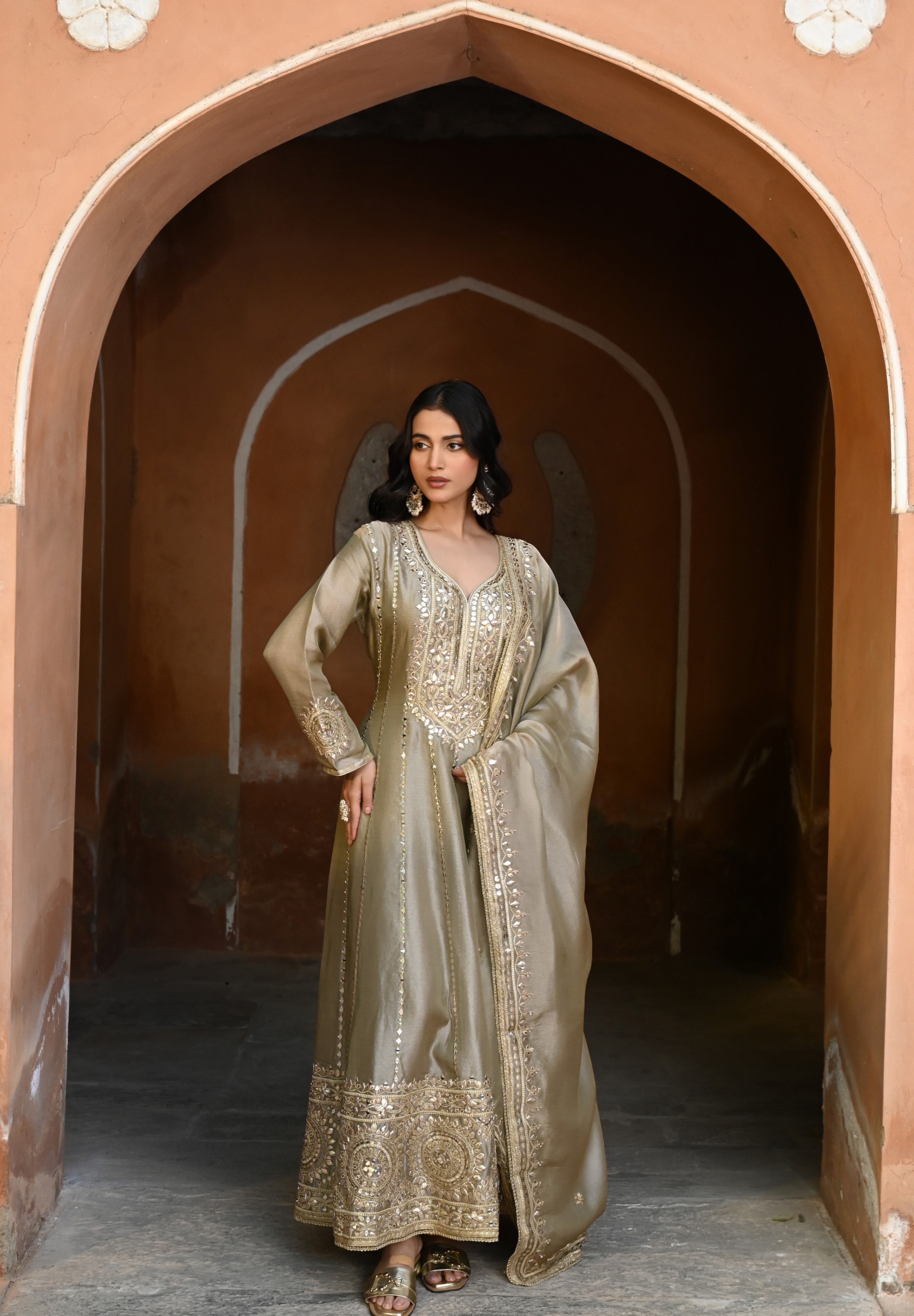 Model wearing an olive-gold embroidered Anarkali dress featuring intricate zari and sequin work, a deep V-neckline, full sleeves, and a rich embellished hem, paired with a matching dupatta and styled under a traditional heritage archway.