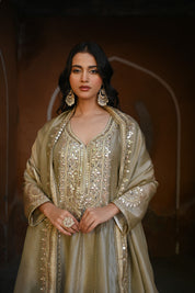 Model wearing an olive-gold embroidered Anarkali dress featuring intricate zari and sequin work, a deep V-neckline, full sleeves, and a rich embellished hem, paired with a matching dupatta and styled under a traditional heritage archway.