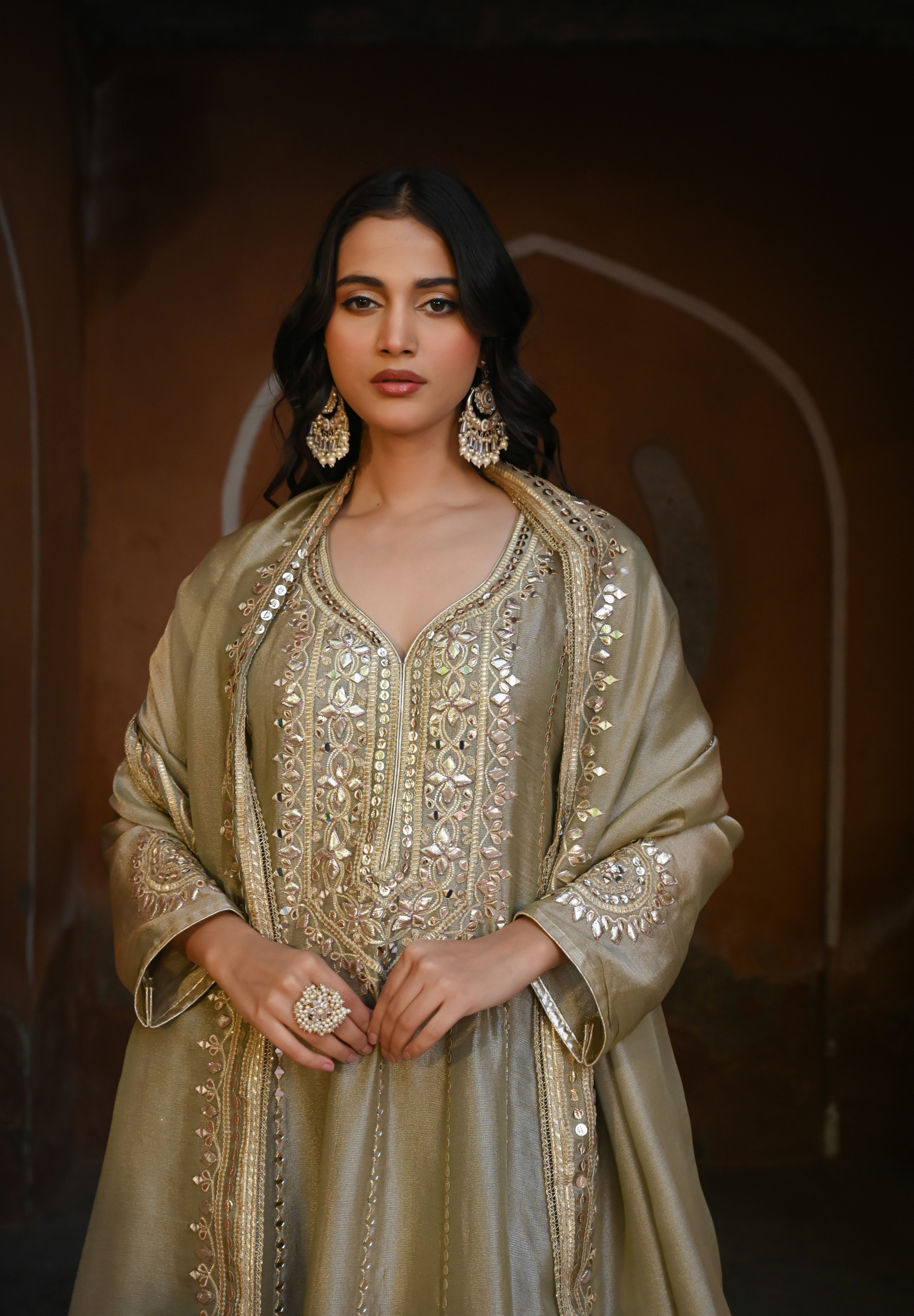 Model wearing an olive-gold embroidered Anarkali dress featuring intricate zari and sequin work, a deep V-neckline, full sleeves, and a rich embellished hem, paired with a matching dupatta and styled under a traditional heritage archway.