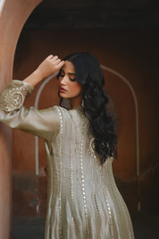 Model wearing an olive-gold embroidered Anarkali dress featuring intricate zari and sequin work, a deep V-neckline, full sleeves, and a rich embellished hem, paired with a matching dupatta and styled under a traditional heritage archway.