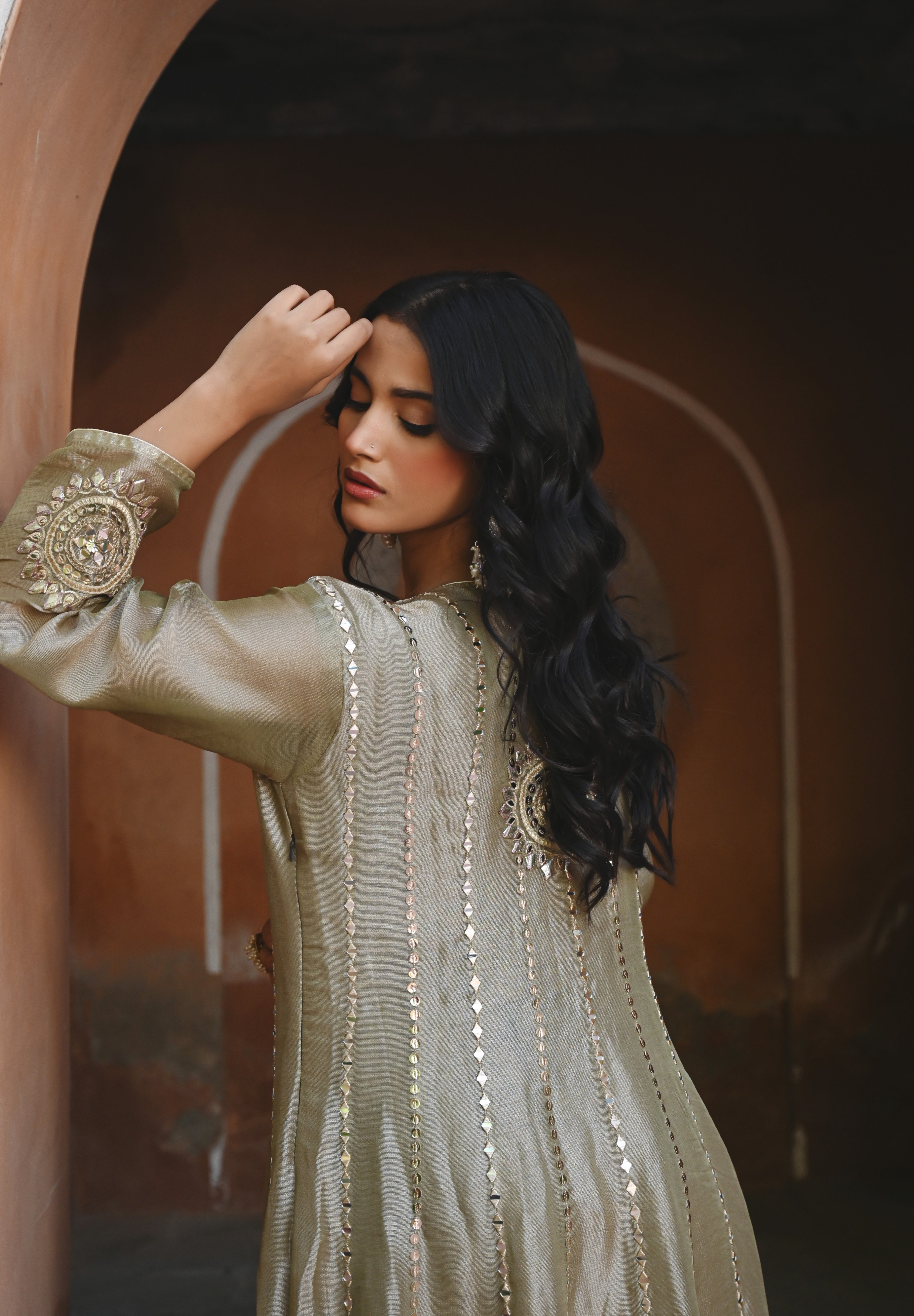 Model wearing an olive-gold embroidered Anarkali dress featuring intricate zari and sequin work, a deep V-neckline, full sleeves, and a rich embellished hem, paired with a matching dupatta and styled under a traditional heritage archway.