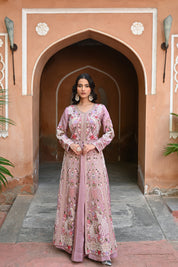 Model wearing a mauve embroidered Anarkali dress with intricate floral and paisley motifs, mirror and thread work detailing, full sleeves, and a flowing floor-length silhouette, styled with statement earrings and standing under a heritage archway.