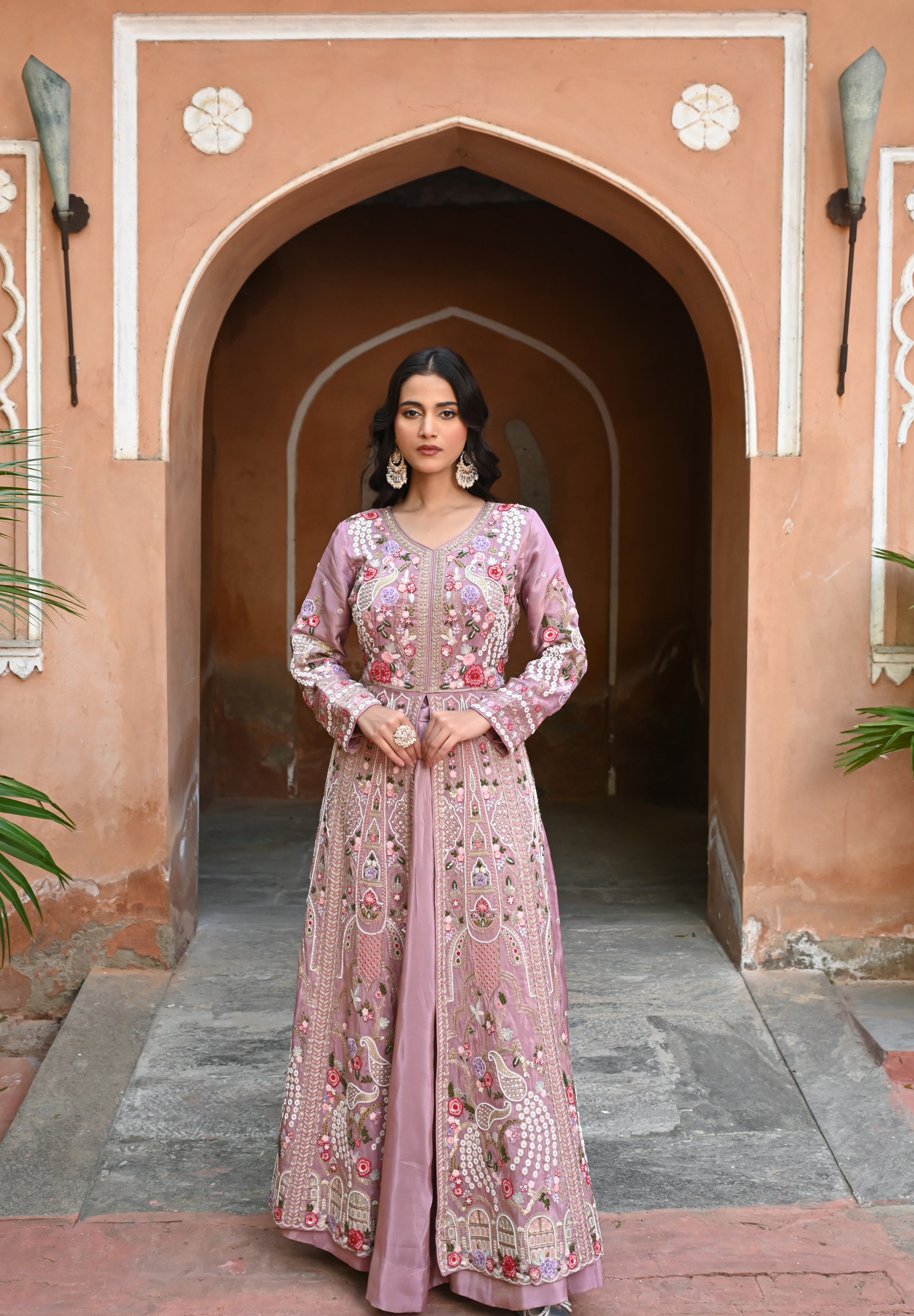 Model wearing a mauve embroidered Anarkali dress with intricate floral and paisley motifs, mirror and thread work detailing, full sleeves, and a flowing floor-length silhouette, styled with statement earrings and standing under a heritage archway.