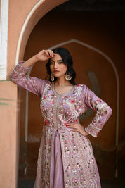 Model wearing a mauve embroidered Anarkali dress with intricate floral and paisley motifs, mirror and thread work detailing, full sleeves, and a flowing floor-length silhouette, styled with statement earrings and standing under a heritage archway.