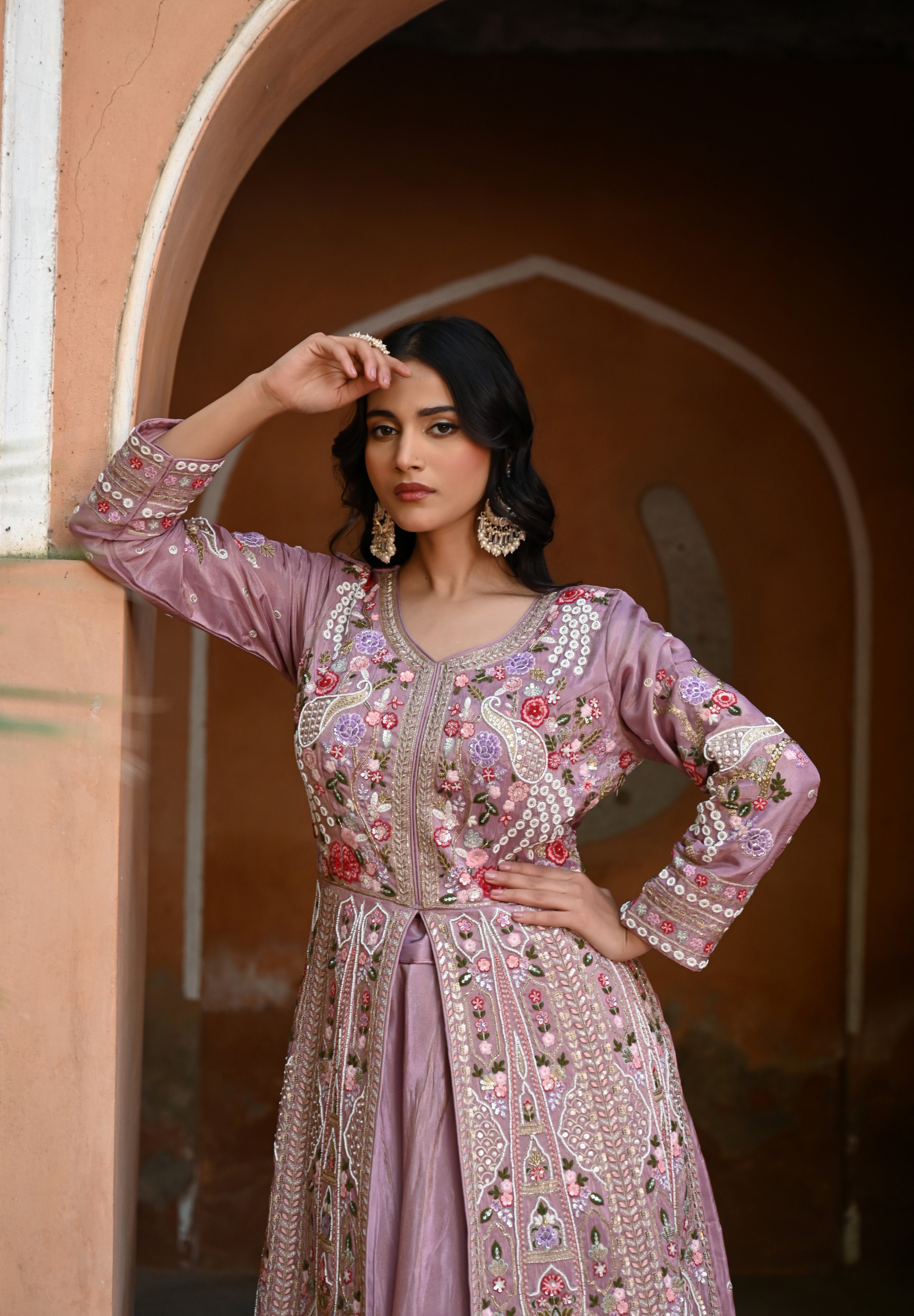 Model wearing a mauve embroidered Anarkali dress with intricate floral and paisley motifs, mirror and thread work detailing, full sleeves, and a flowing floor-length silhouette, styled with statement earrings and standing under a heritage archway.