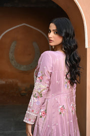 Model wearing a mauve embroidered Anarkali dress with intricate floral and paisley motifs, mirror and thread work detailing, full sleeves, and a flowing floor-length silhouette, styled with statement earrings and standing under a heritage archway.