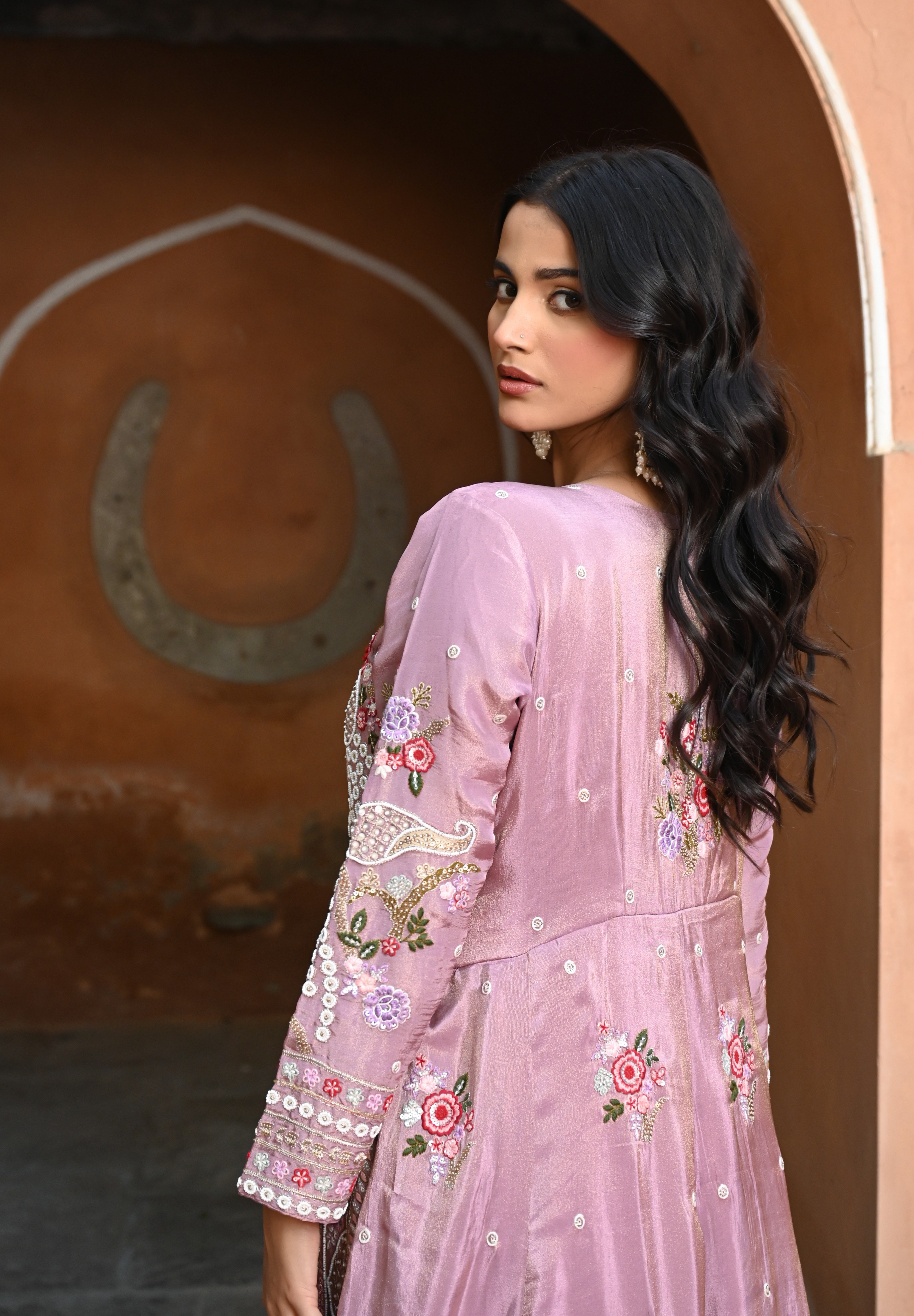 Model wearing a mauve embroidered Anarkali dress with intricate floral and paisley motifs, mirror and thread work detailing, full sleeves, and a flowing floor-length silhouette, styled with statement earrings and standing under a heritage archway.