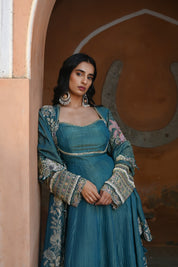 Model wearing a teal blue Anarkali dress with a fitted bodice, square neckline, and full sleeves adorned with intricate floral embroidery and gold accents, featuring a softly flared silhouette, styled with statement earrings against a heritage archway backdrop.