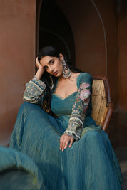 Model wearing a teal blue Anarkali dress with a fitted bodice, square neckline, and full sleeves adorned with intricate floral embroidery and gold accents, featuring a softly flared silhouette, styled with statement earrings against a heritage archway backdrop.