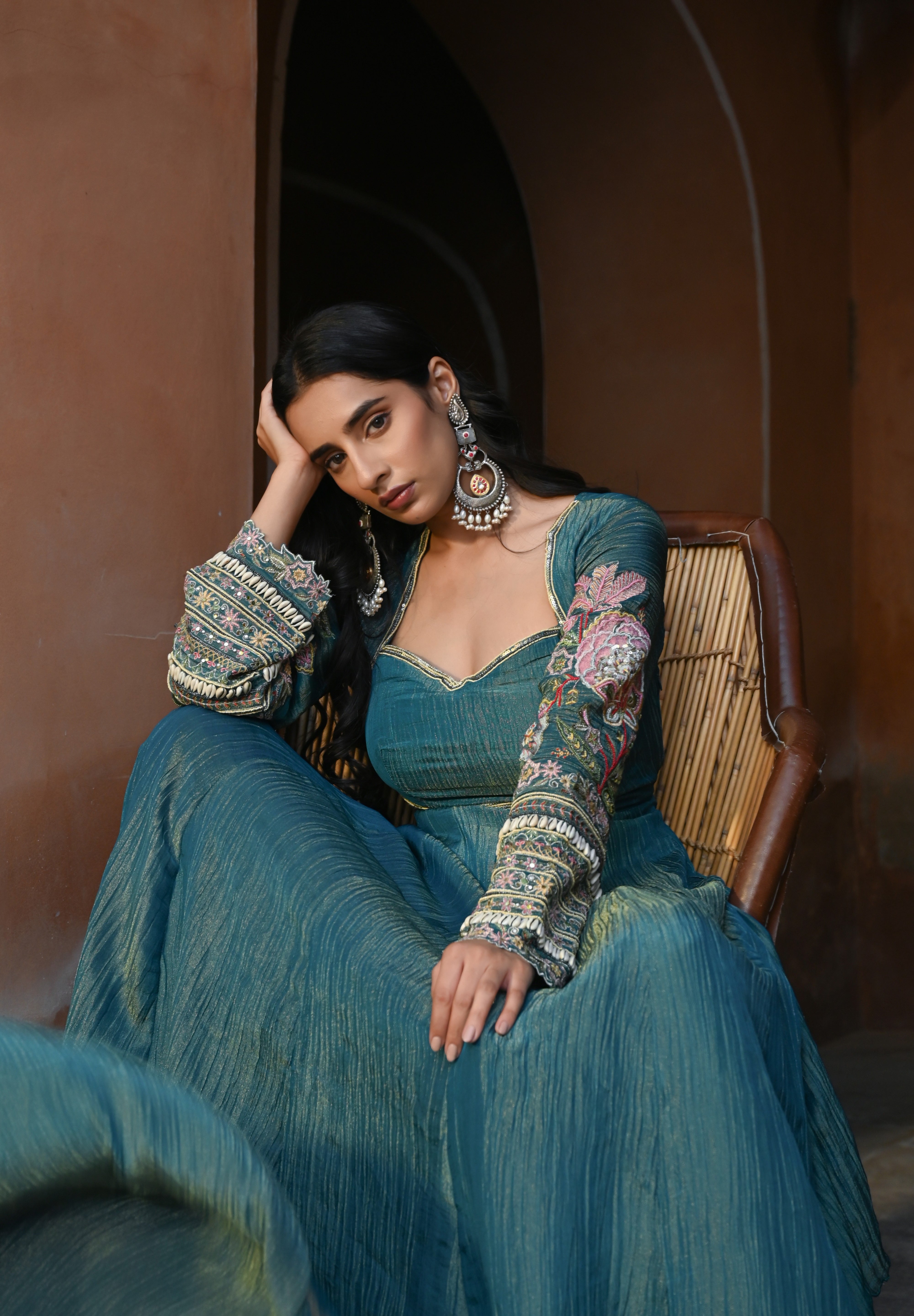 Model wearing a teal blue Anarkali dress with a fitted bodice, square neckline, and full sleeves adorned with intricate floral embroidery and gold accents, featuring a softly flared silhouette, styled with statement earrings against a heritage archway backdrop.