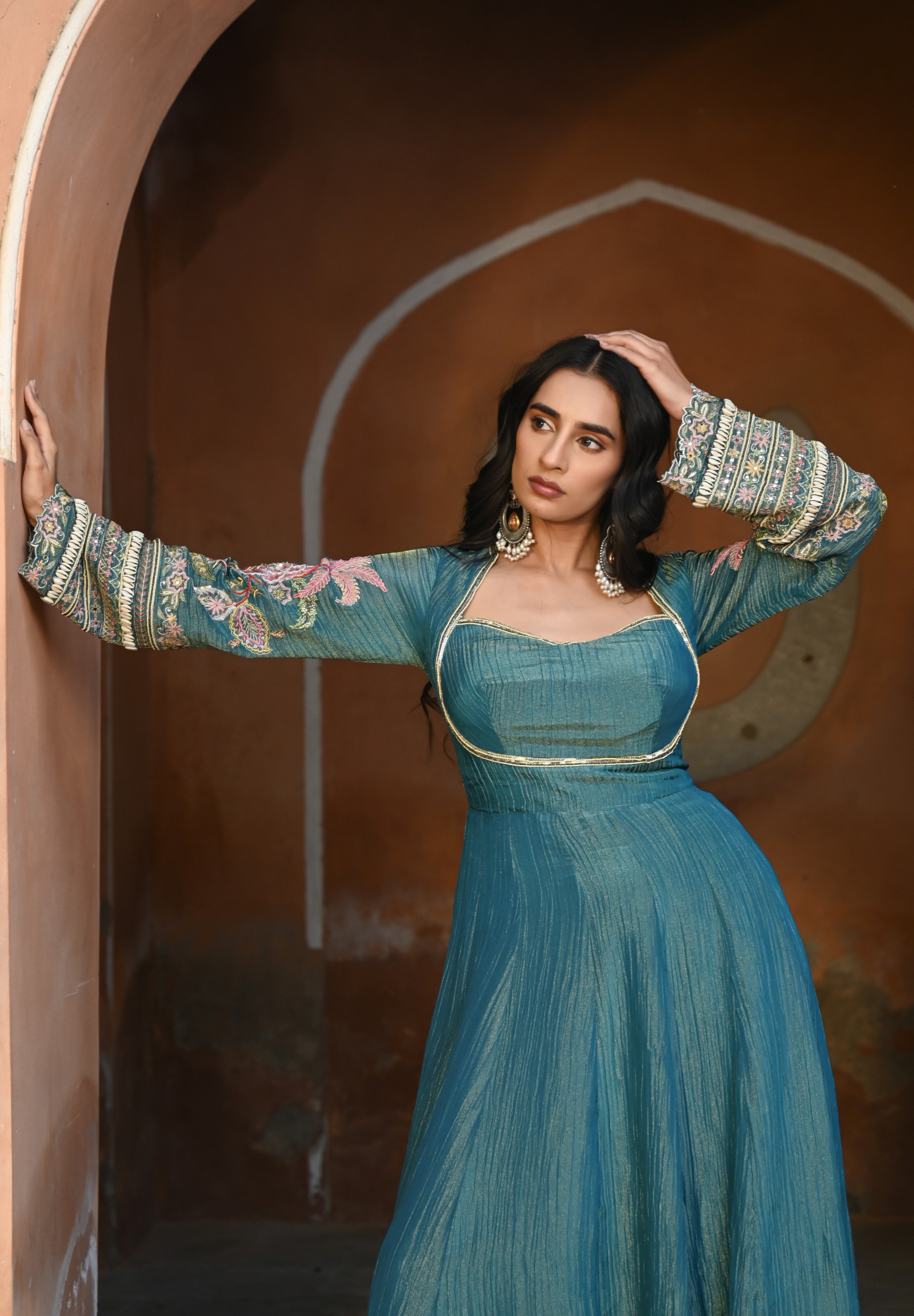 Model wearing a teal blue Anarkali dress with a fitted bodice, square neckline, and full sleeves adorned with intricate floral embroidery and gold accents, featuring a softly flared silhouette, styled with statement earrings against a heritage archway backdrop.