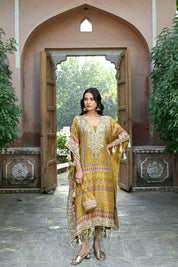 Model wearing a mustard yellow embroidered kaftan-style dress featuring intricate mirror work, geometric and floral motifs, tassel detailing on the sleeves and hem, and a relaxed silhouette, styled with gold footwear and statement earrings in a heritage courtyard setting.