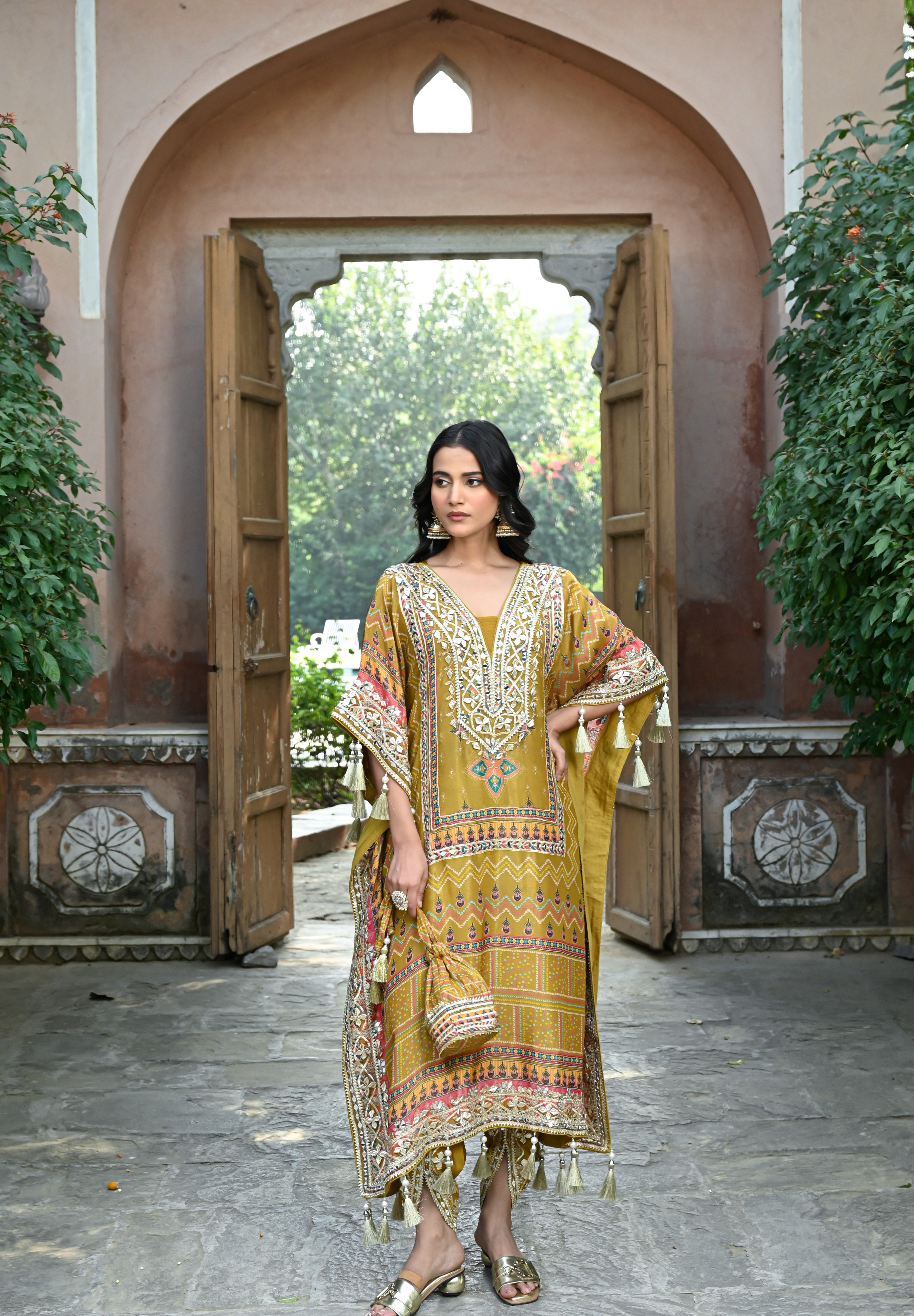 Model wearing a mustard yellow embroidered kaftan-style dress featuring intricate mirror work, geometric and floral motifs, tassel detailing on the sleeves and hem, and a relaxed silhouette, styled with gold footwear and statement earrings in a heritage courtyard setting.