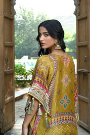 Model wearing a mustard yellow embroidered kaftan-style dress featuring intricate mirror work, geometric and floral motifs, tassel detailing on the sleeves and hem, and a relaxed silhouette, styled with gold footwear and statement earrings in a heritage courtyard setting.