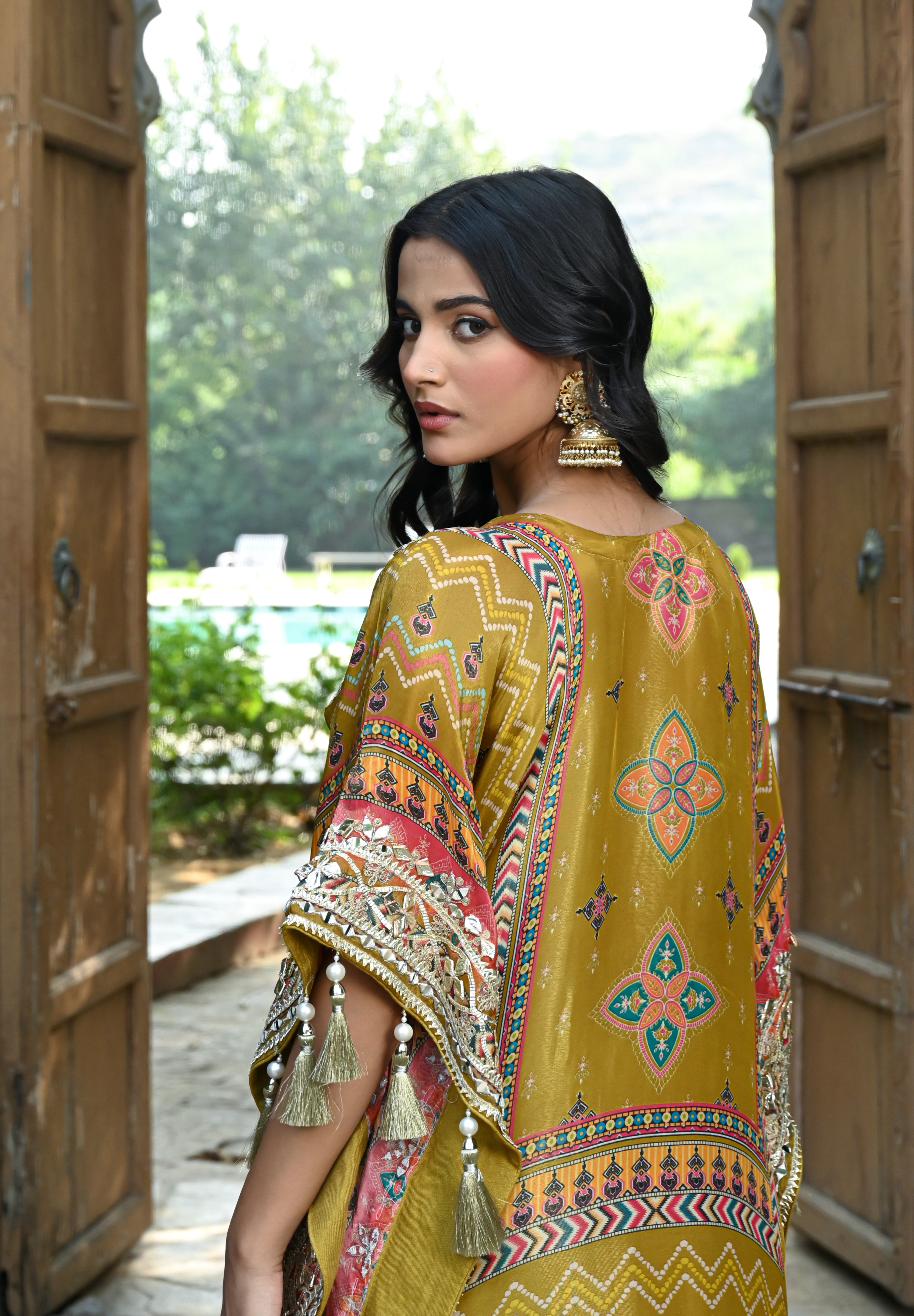 Model wearing a mustard yellow embroidered kaftan-style dress featuring intricate mirror work, geometric and floral motifs, tassel detailing on the sleeves and hem, and a relaxed silhouette, styled with gold footwear and statement earrings in a heritage courtyard setting.