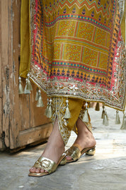 Model wearing a mustard yellow embroidered kaftan-style dress featuring intricate mirror work, geometric and floral motifs, tassel detailing on the sleeves and hem, and a relaxed silhouette, styled with gold footwear and statement earrings in a heritage courtyard setting.