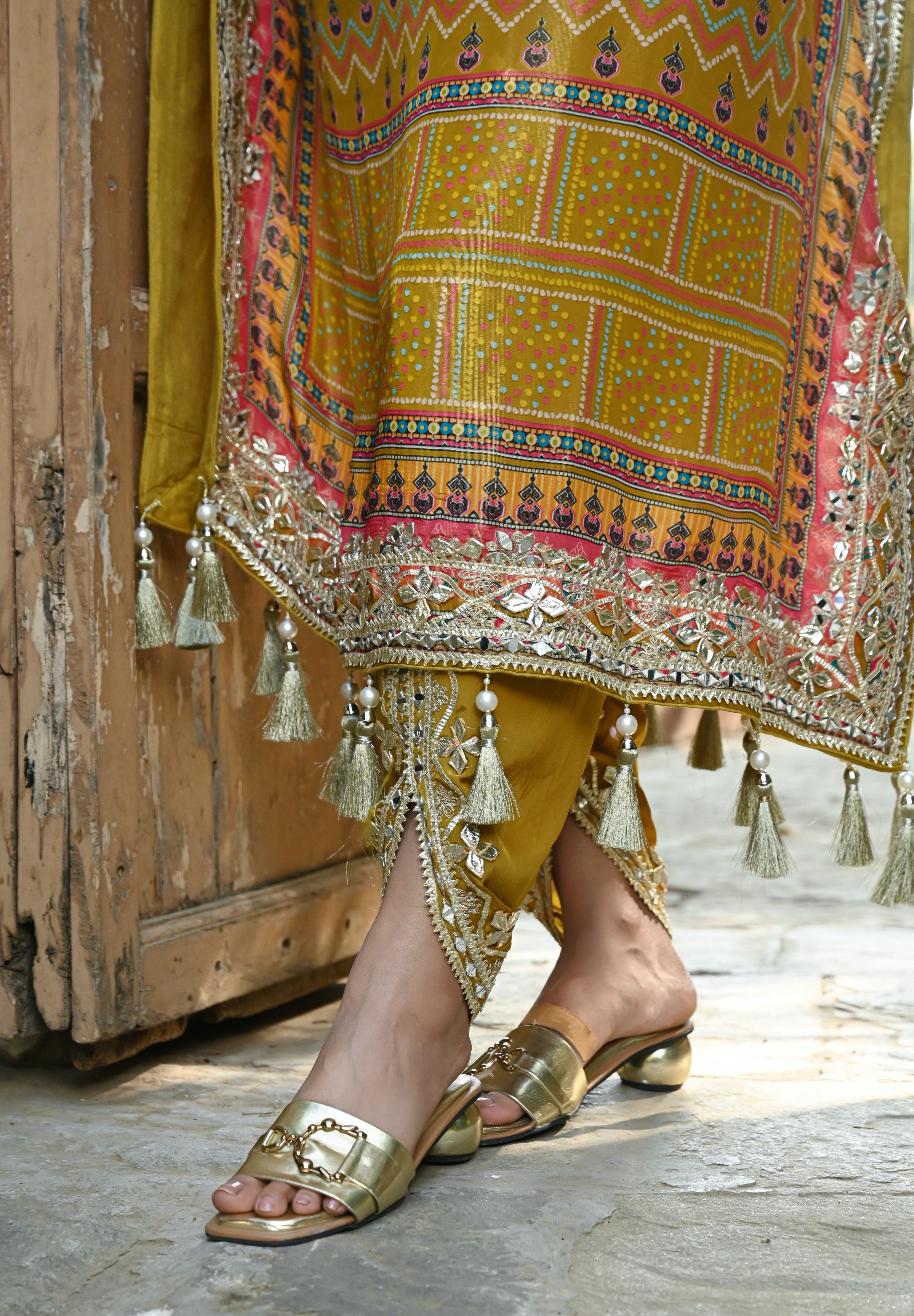 Model wearing a mustard yellow embroidered kaftan-style dress featuring intricate mirror work, geometric and floral motifs, tassel detailing on the sleeves and hem, and a relaxed silhouette, styled with gold footwear and statement earrings in a heritage courtyard setting.
