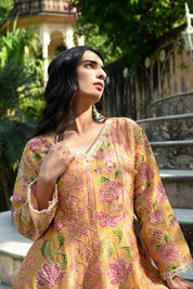 Model wearing a mustard yellow embroidered kurta set featuring intricate floral motifs in pink and green, delicate mirror and thread work, full sleeves, and a straight silhouette, paired with matching pants and a flowing floral dupatta, styled in an outdoor heritage garden setting.