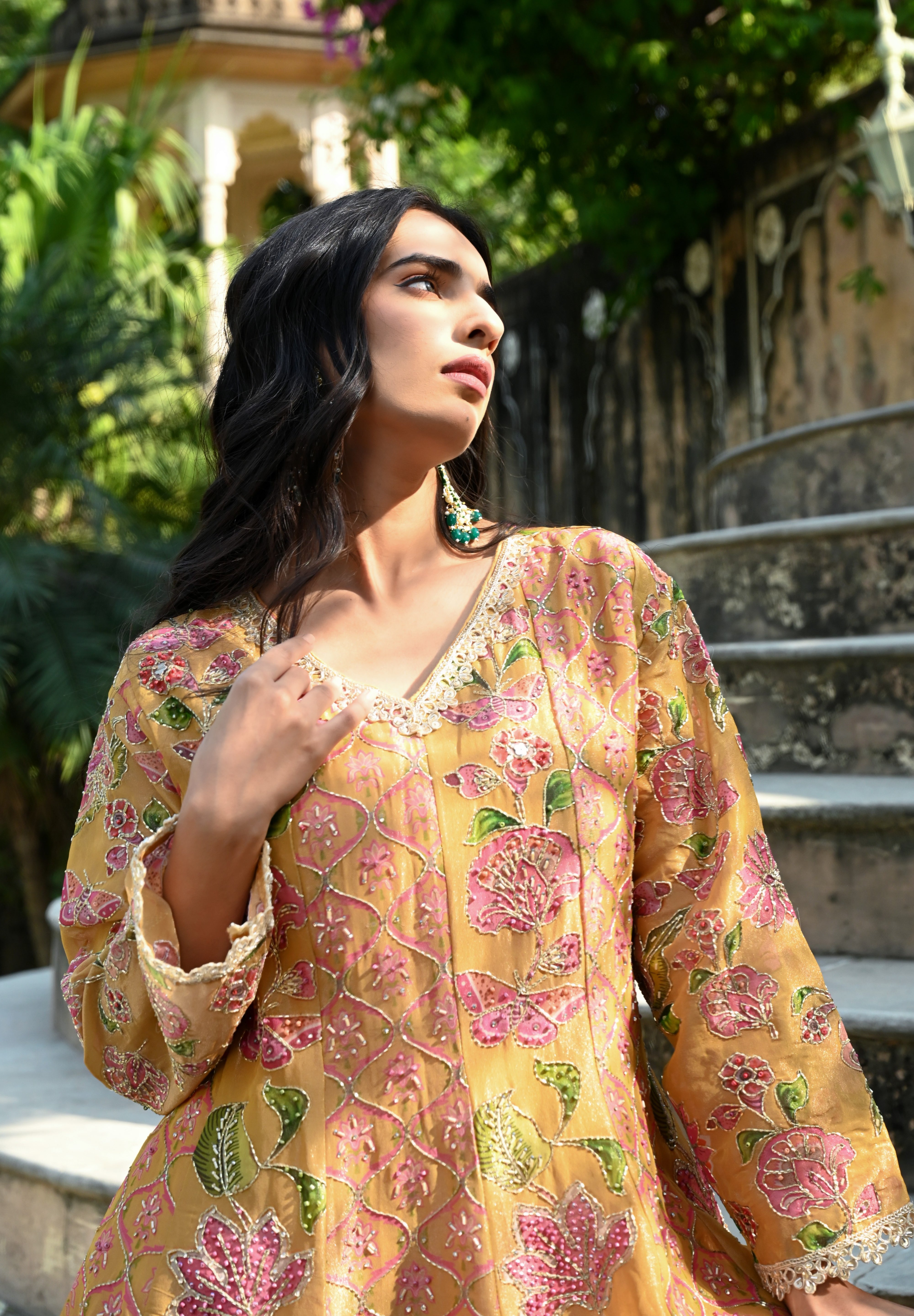 Model wearing a mustard yellow embroidered kurta set featuring intricate floral motifs in pink and green, delicate mirror and thread work, full sleeves, and a straight silhouette, paired with matching pants and a flowing floral dupatta, styled in an outdoor heritage garden setting.